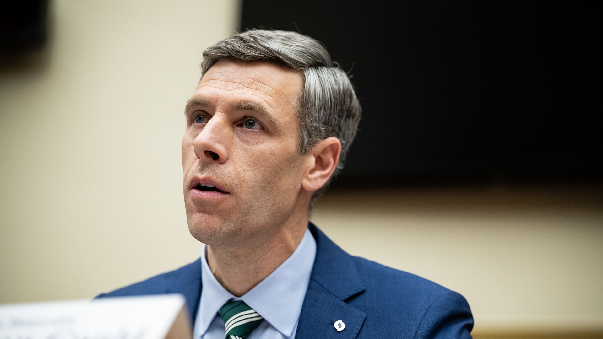 Man with short grey hair wearing a blue suit, light blue shirt, and green striped tie speaking at a microphone, with a blurred nameplate in front of him against a beige and black background.