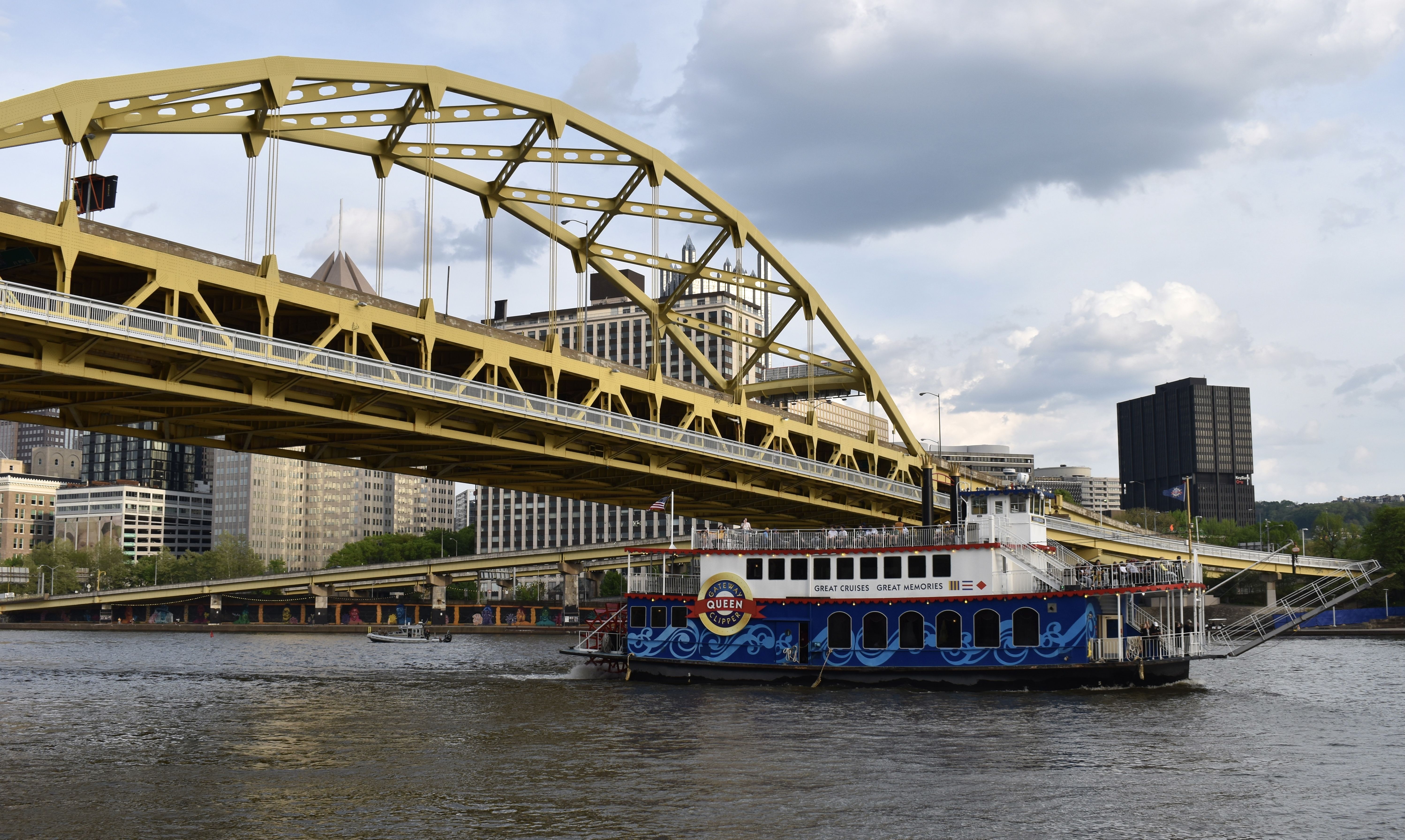 A large yellow steel arch bridge spans a brown river, with a blue and white passenger boat named Queen docked beneath. City buildings and a cloudy sky form the backdrop.