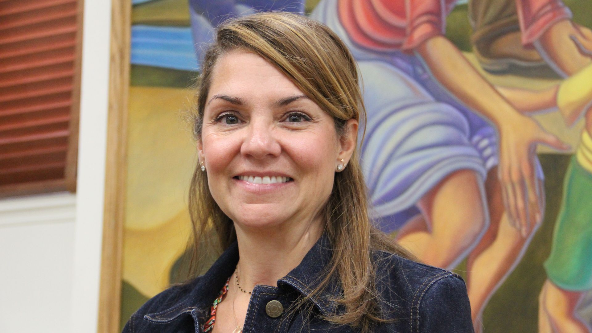 A woman smiles while wearing a dark denim jacket and standing in front of an oil painting.