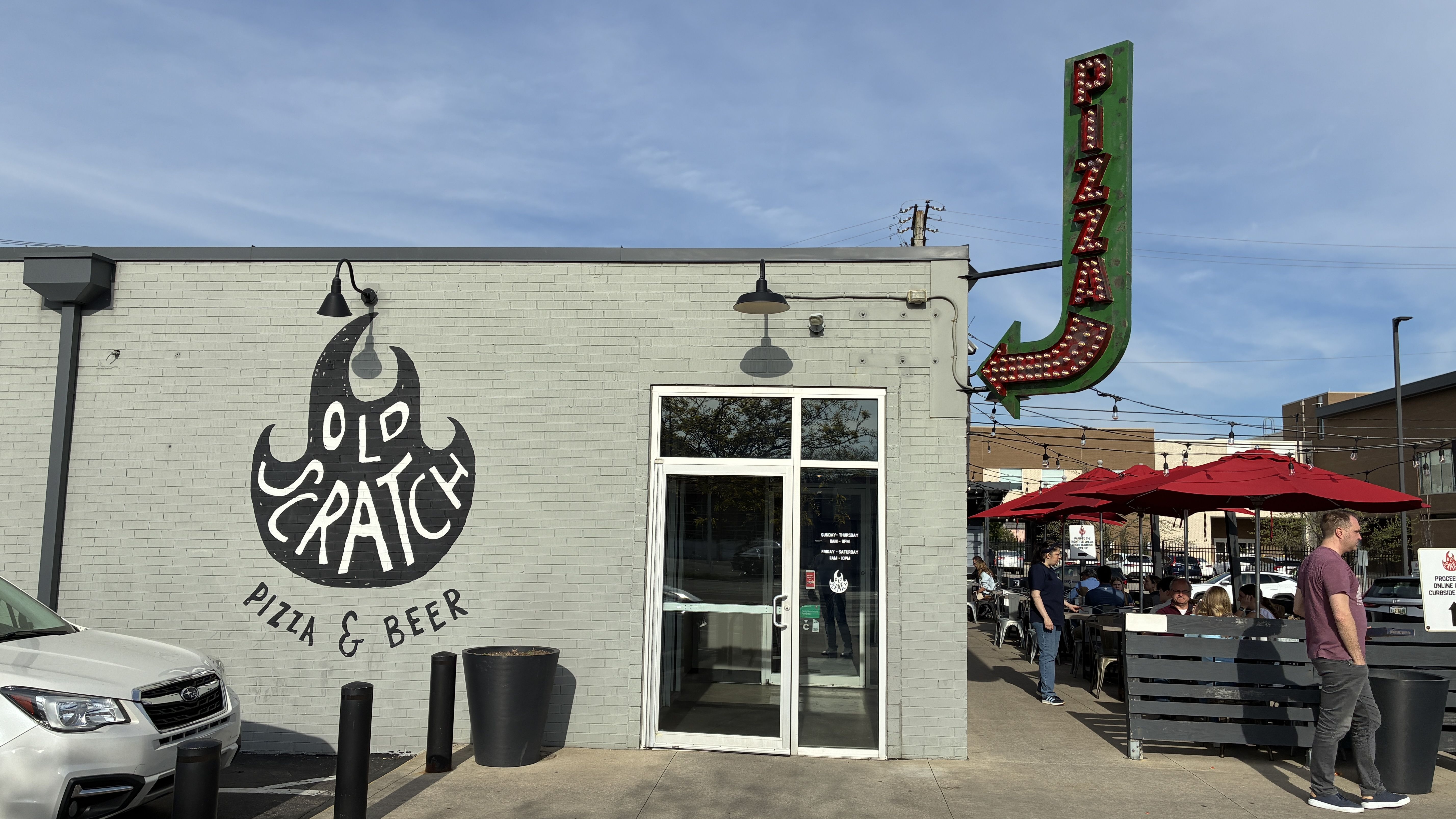 Light gray-green brick pizza and beer shop exterior with a tall green neon PIZZA sign. A circular logo on the wall reads Old Scratch Pizza & Beer. Outdoor seating under red umbrellas; a white car is parked outside.