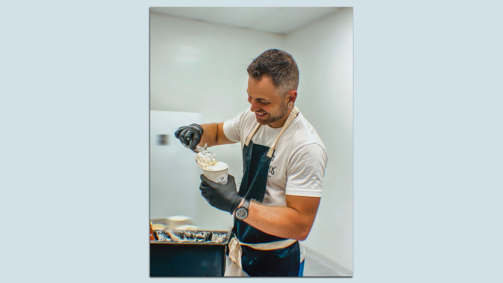 Smiling man in a white t-shirt and dark apron with beige straps, wearing black gloves, scooping ice cream into a cup with a metal scoop in a bright, clean kitchen.
