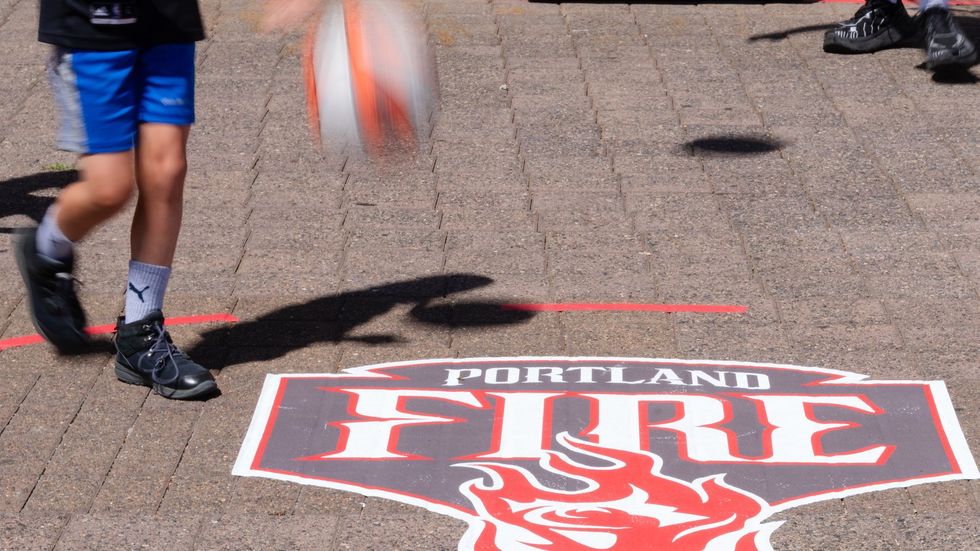 Blurry orange-and-white basketball moves across a brick-paved court as a player in blue shorts and black sneakers passes by; a large Portland Fire sign with a red flame lies on the ground.