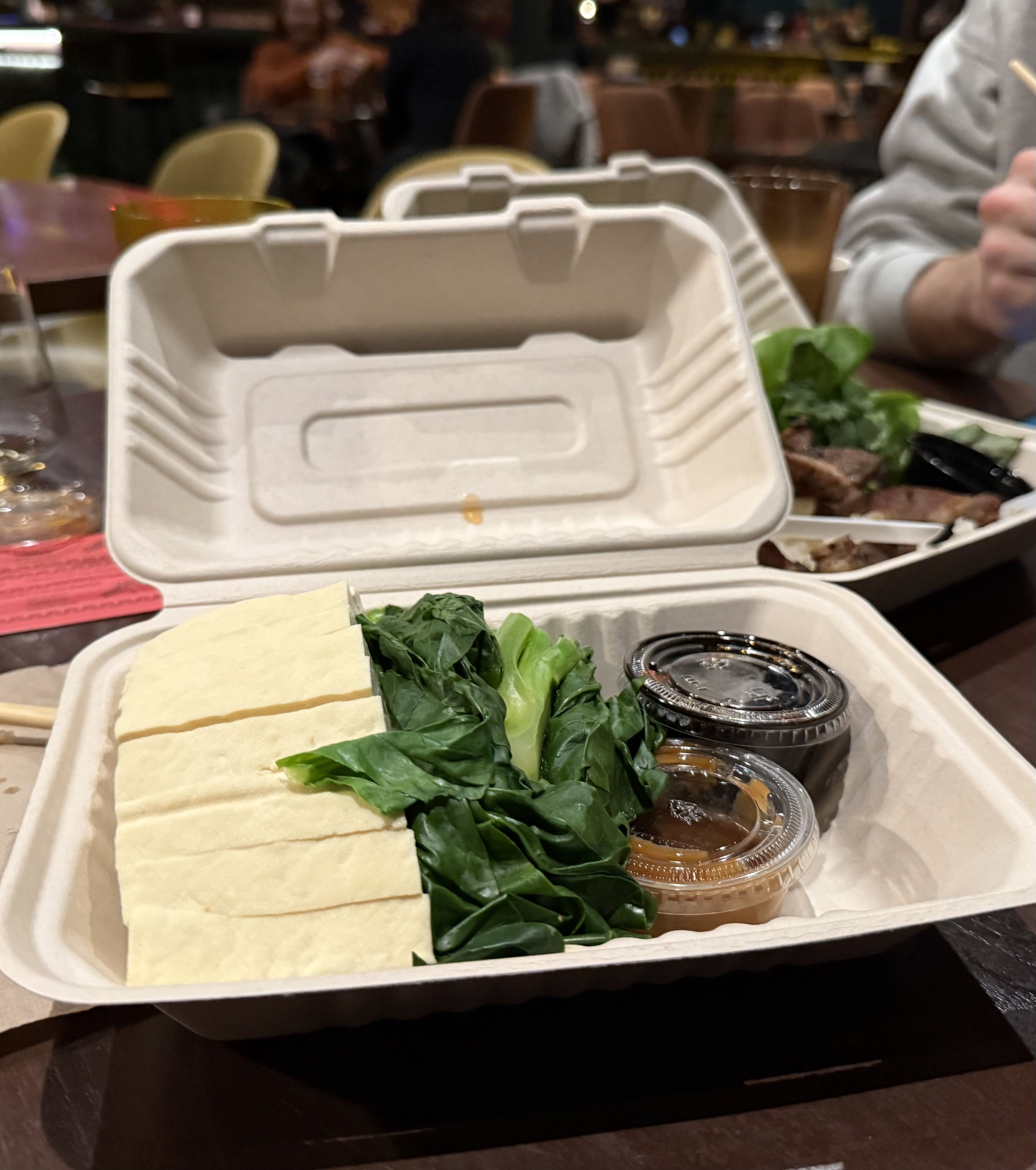 Open beige takeout container with sliced tofu, green leafy vegetables, and two small plastic sauce containers, on a dark table in a dimly lit restaurant.
