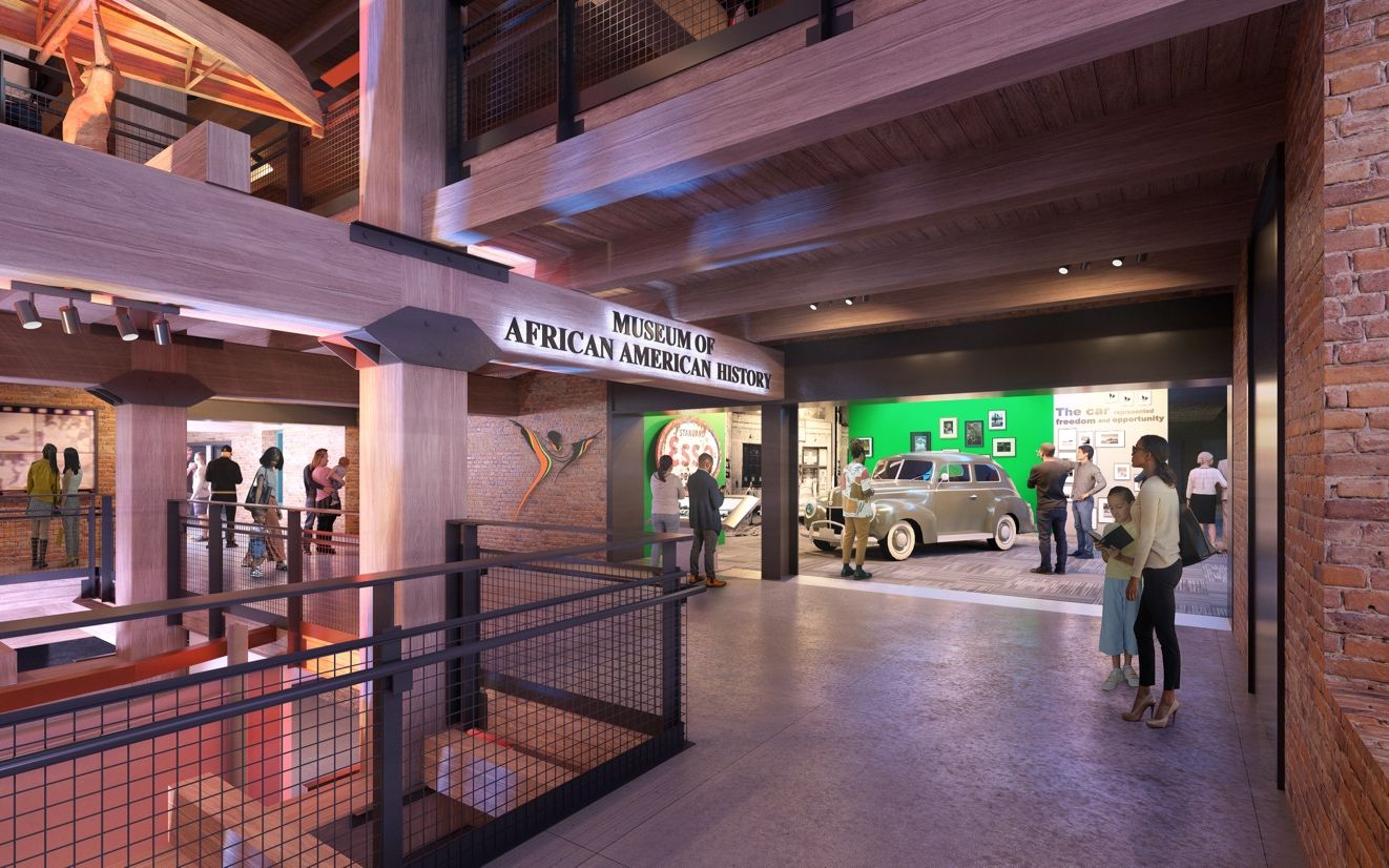Interior of a two-level museum with brick walls and timber beams. A large sign says "Museum of African American History" while visitors explore a vintage car exhibit and displays.