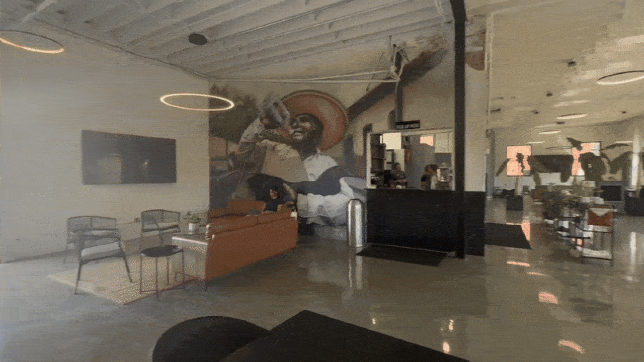 Modern office lobby with polished floors and ceiling lights, a large mural of a man drinking from a bottle, orange couch, black chairs, and an open office area in the background.