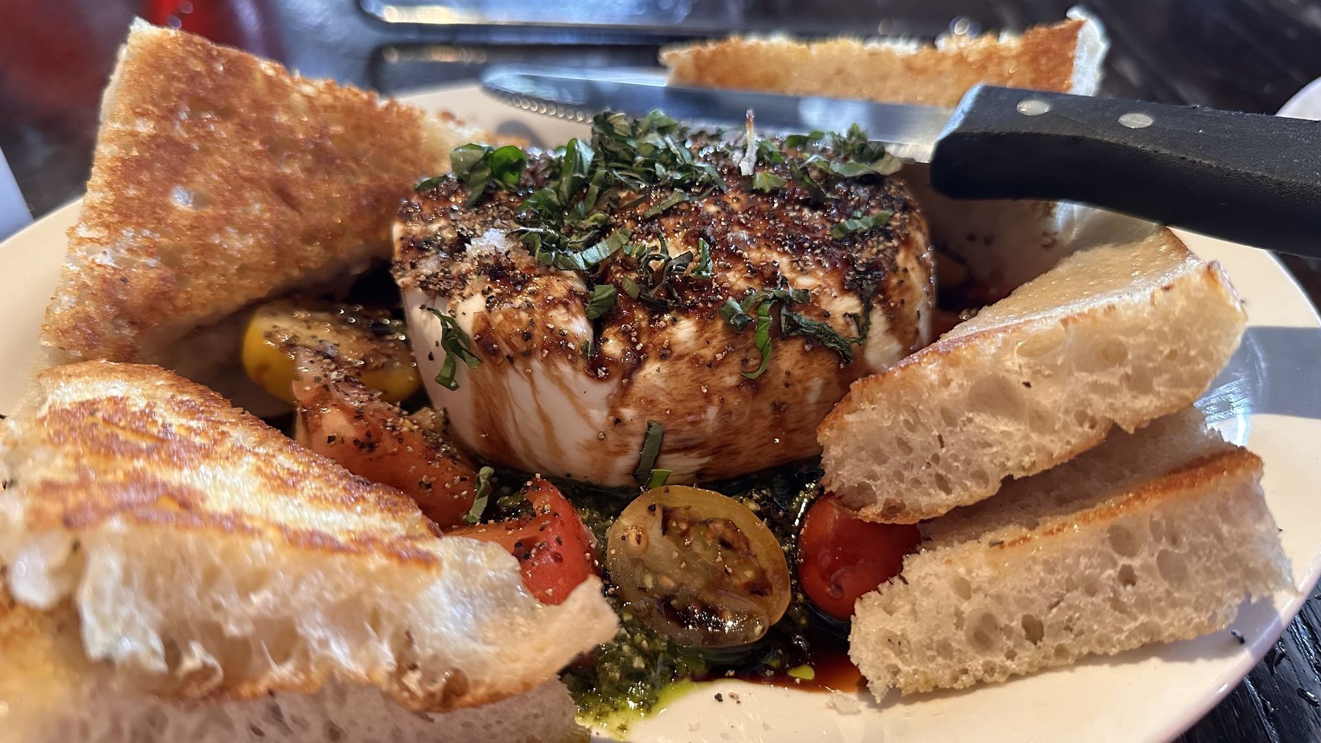 Plate with burrata topped with herbs and seasoning, surrounded by toasted bread and cherry tomatoes, with a knife resting on the plate.