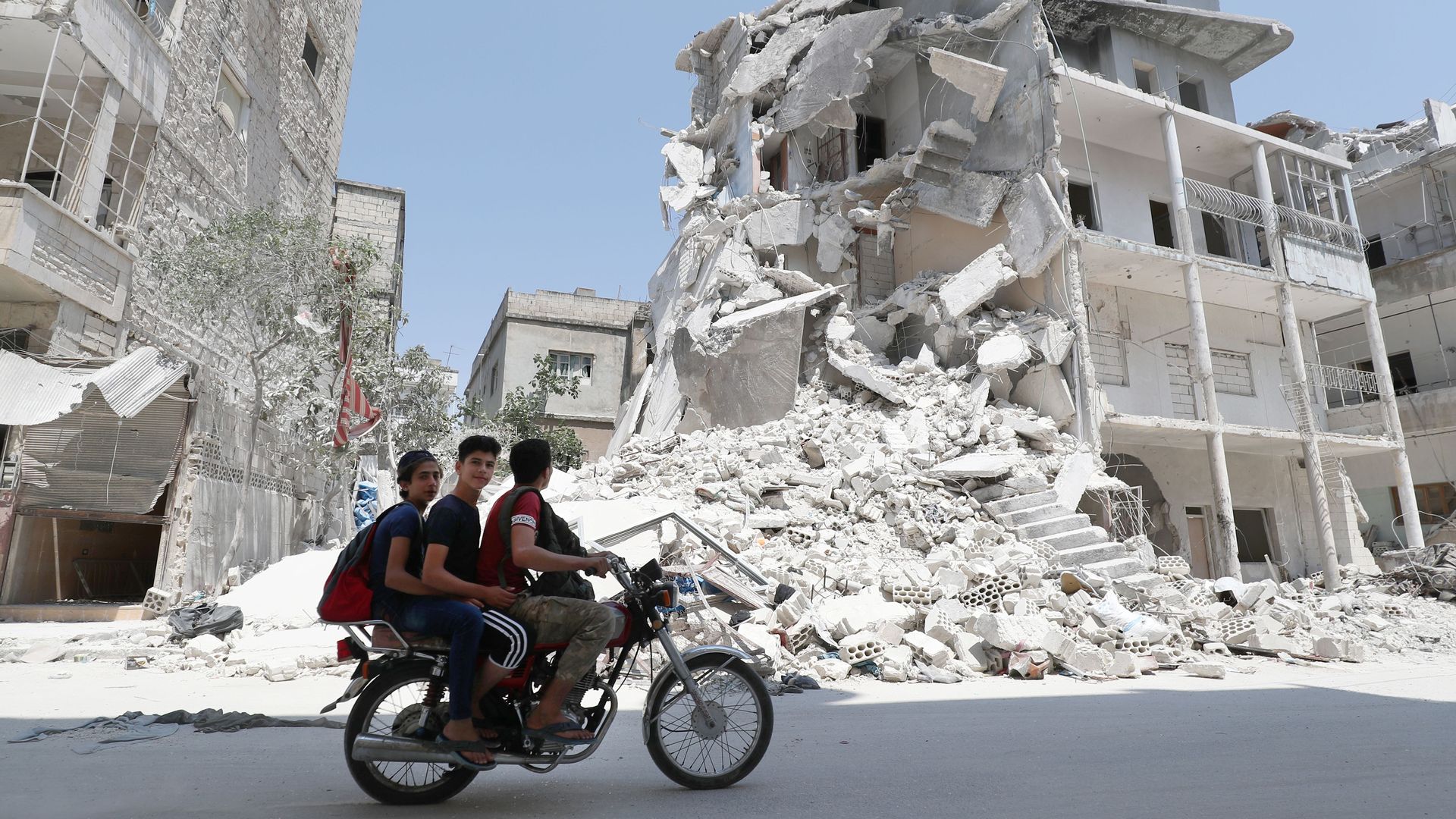 Men on a motorcycle ride by an airstrike-damaged building