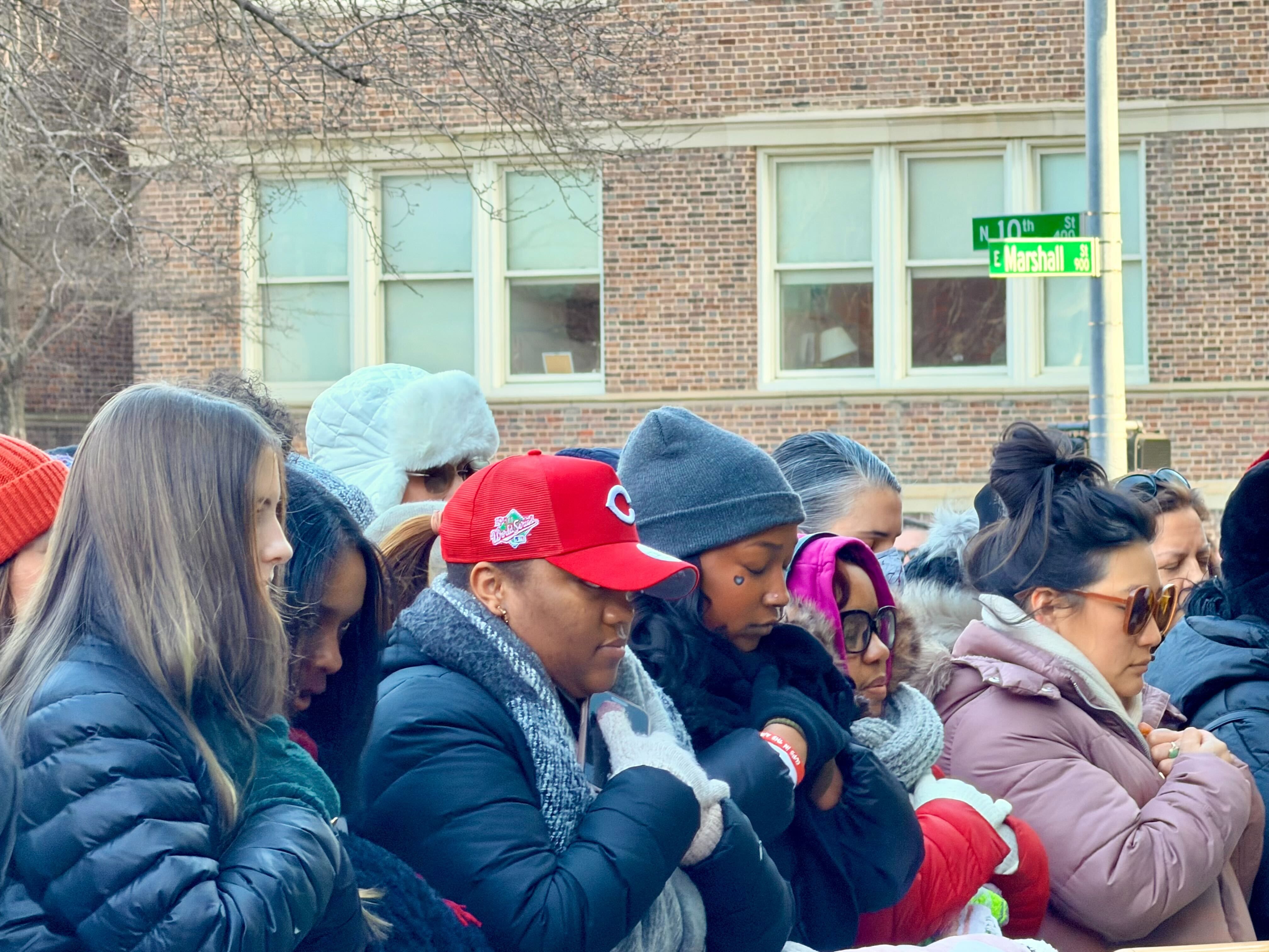 A group of people bundled in winter clothing stand closely together outdoors near a brick building by street signs reading N 10th St and E Marshall St.