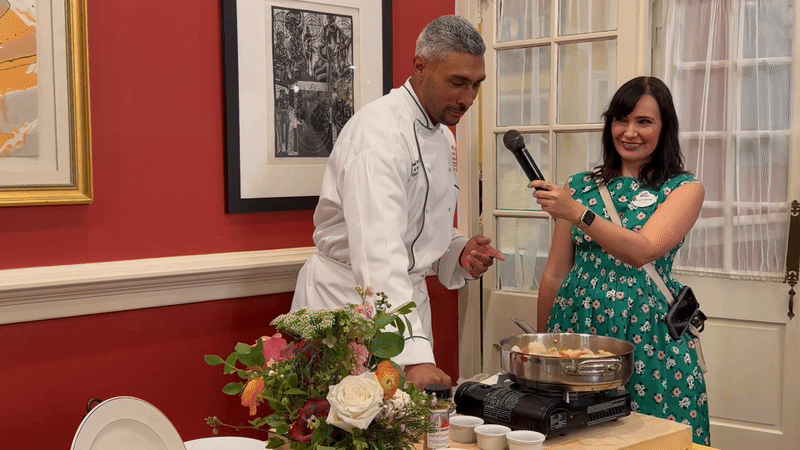 Chef in white coat cooking on portable stove as woman in green floral dress holds microphone, standing in red-walled room with framed art and flower centerpiece on table.