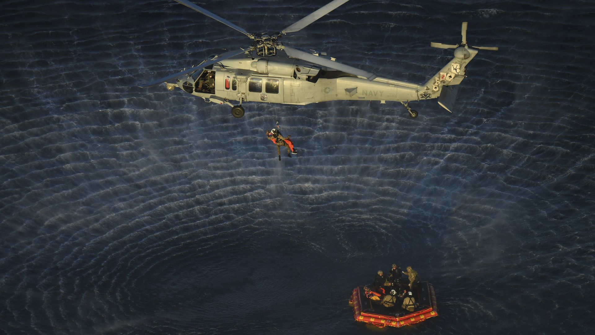 A U.S. Navy helicopter hovers over the ocean, lowering a rescuer by cable toward a small inflatable raft below, where several people in life vests wait amid rippling water.
