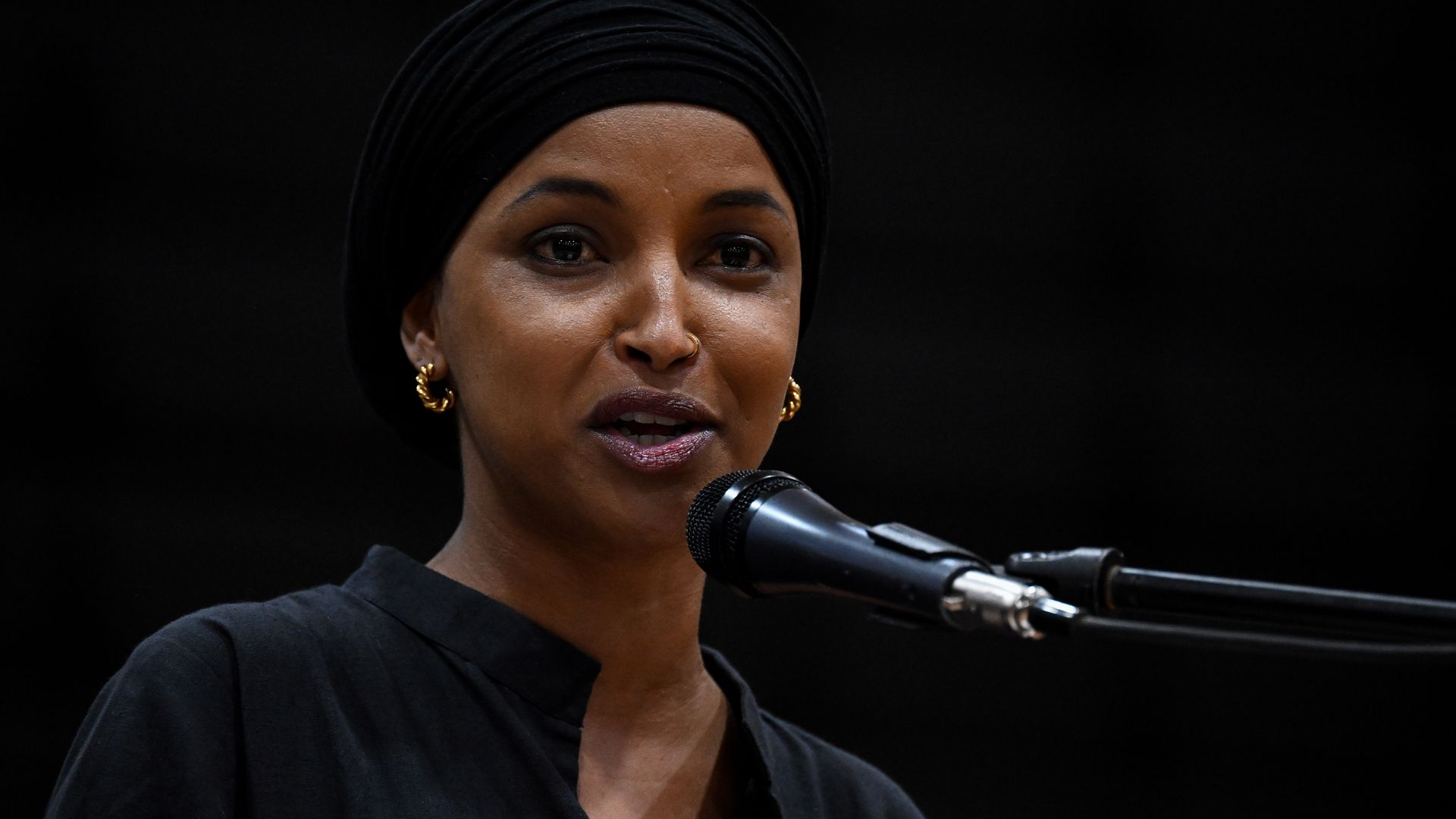 Rep. Ilhan Omar speaks at a microphone against a black background.