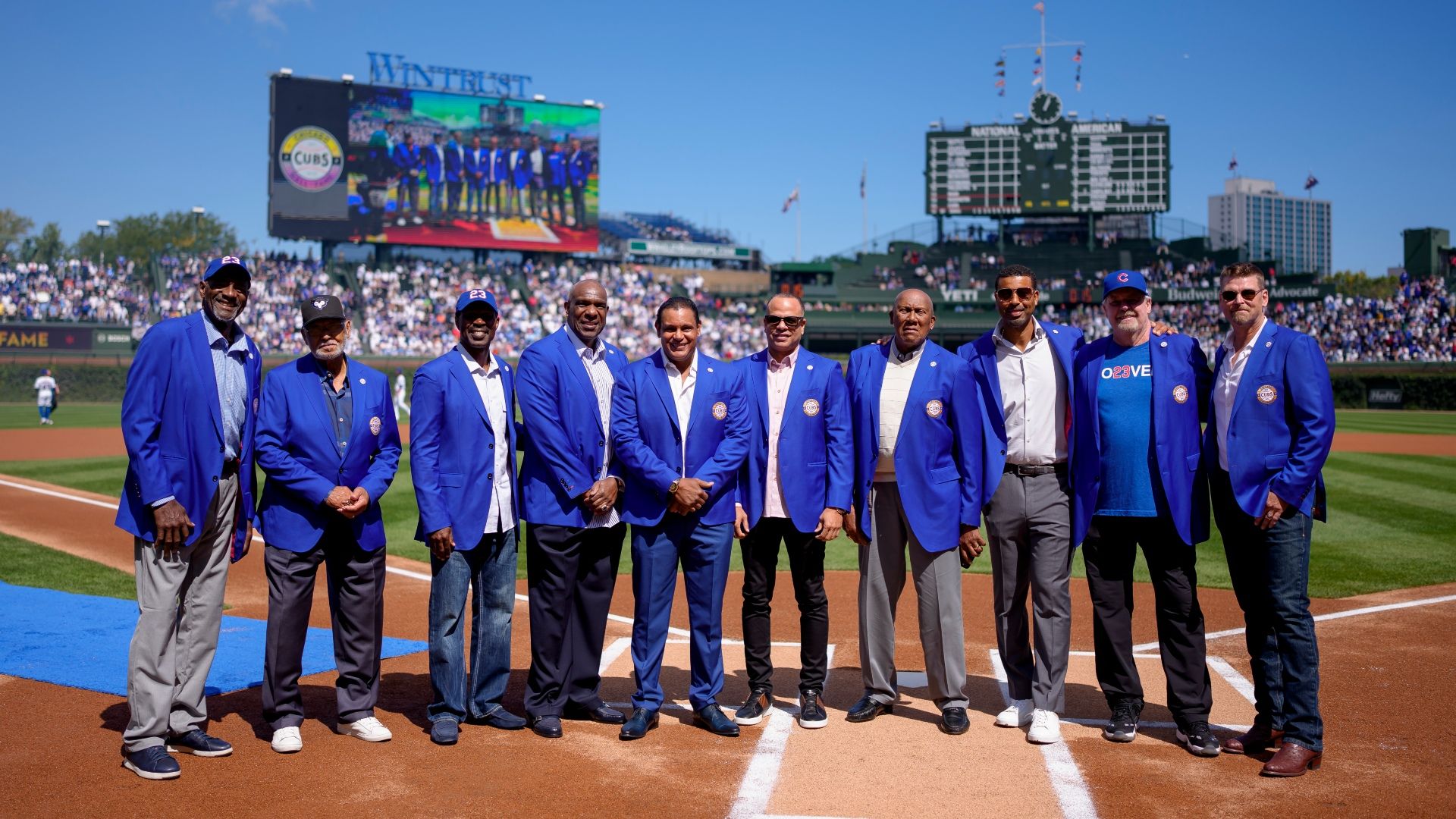 Group of men in matching blue blazers posing on a baseball field, with a large scoreboard and video screen behind them and a crowded stadium under a clear blue sky.