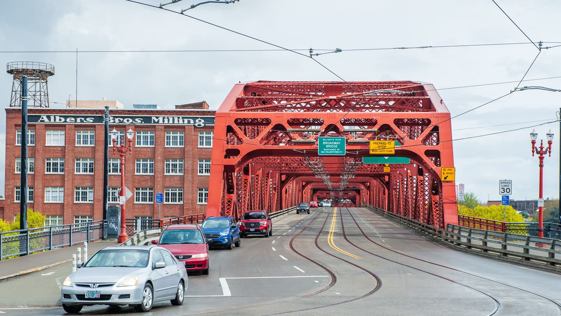 Red steel Broadway Bridge with cars driving and parked, old brick Albers Bros. Milling building on left, cloudy sky and street signs including speed limit 30 and bridge height limit.