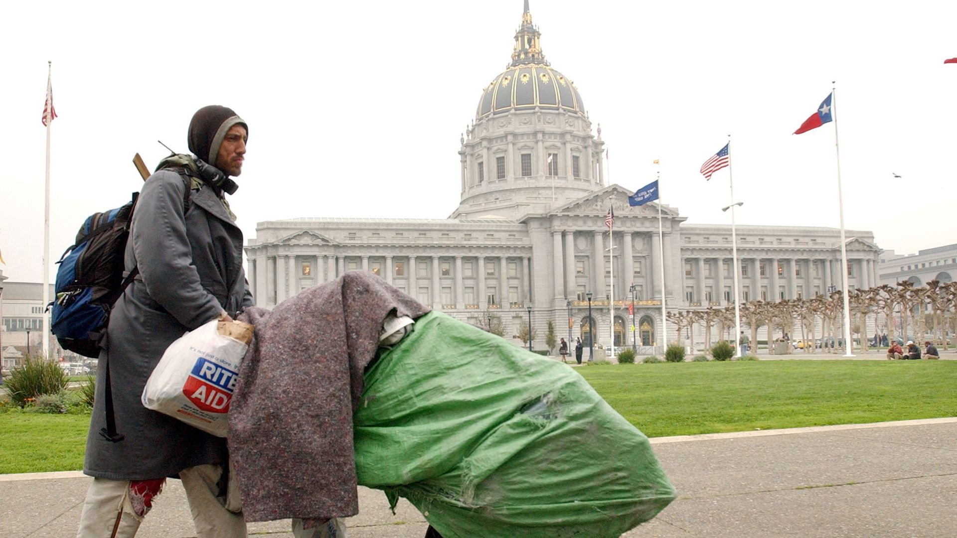 Homeless man pushes green rolly bag in front of San Francisco City Hall dome.