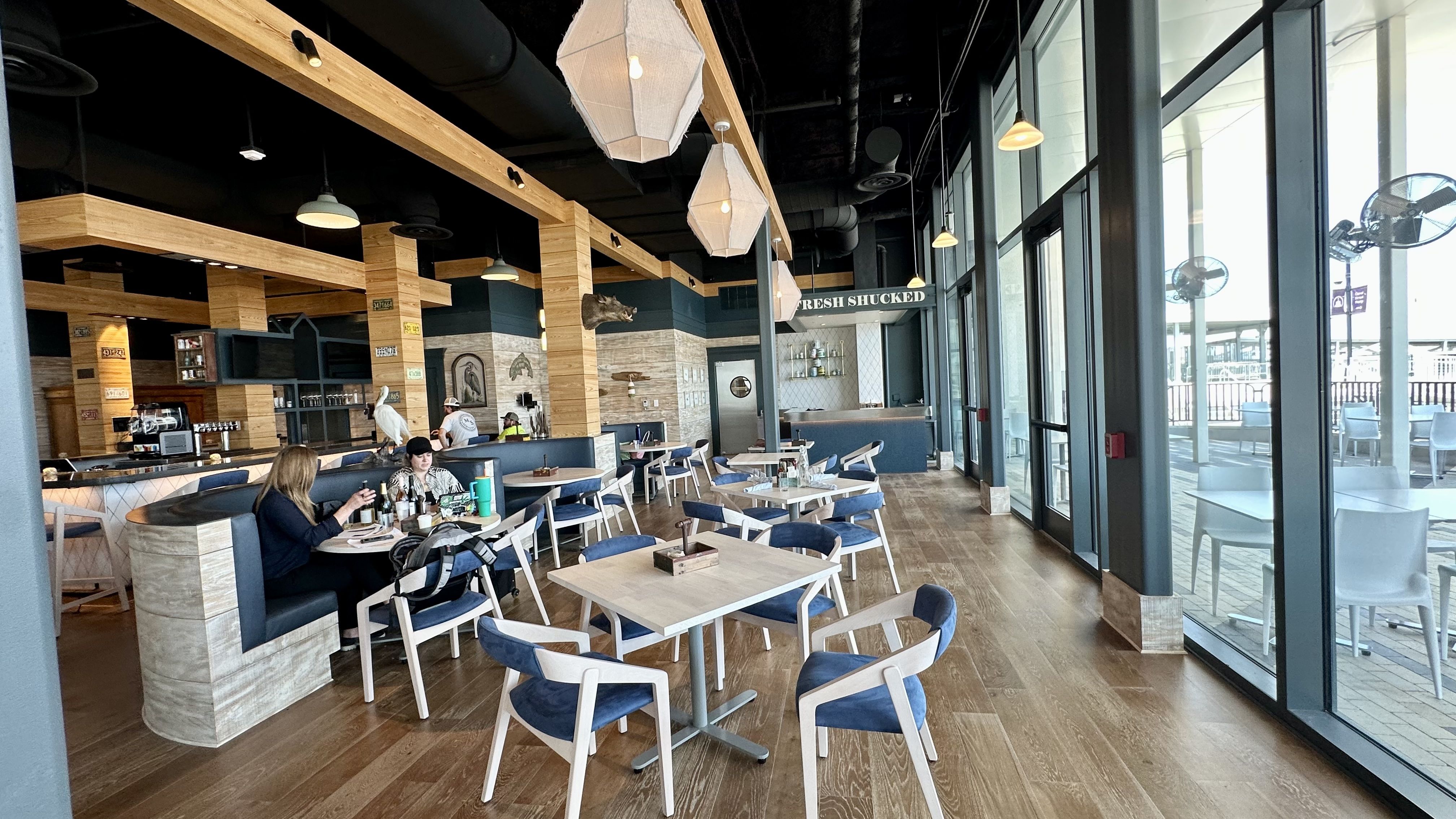 Modern restaurant interior with wood floors, blue cushioned chairs, wooden tables, large windows on the right, pendant lights, and a "FRESH SHUCKED" sign above the bar area.
