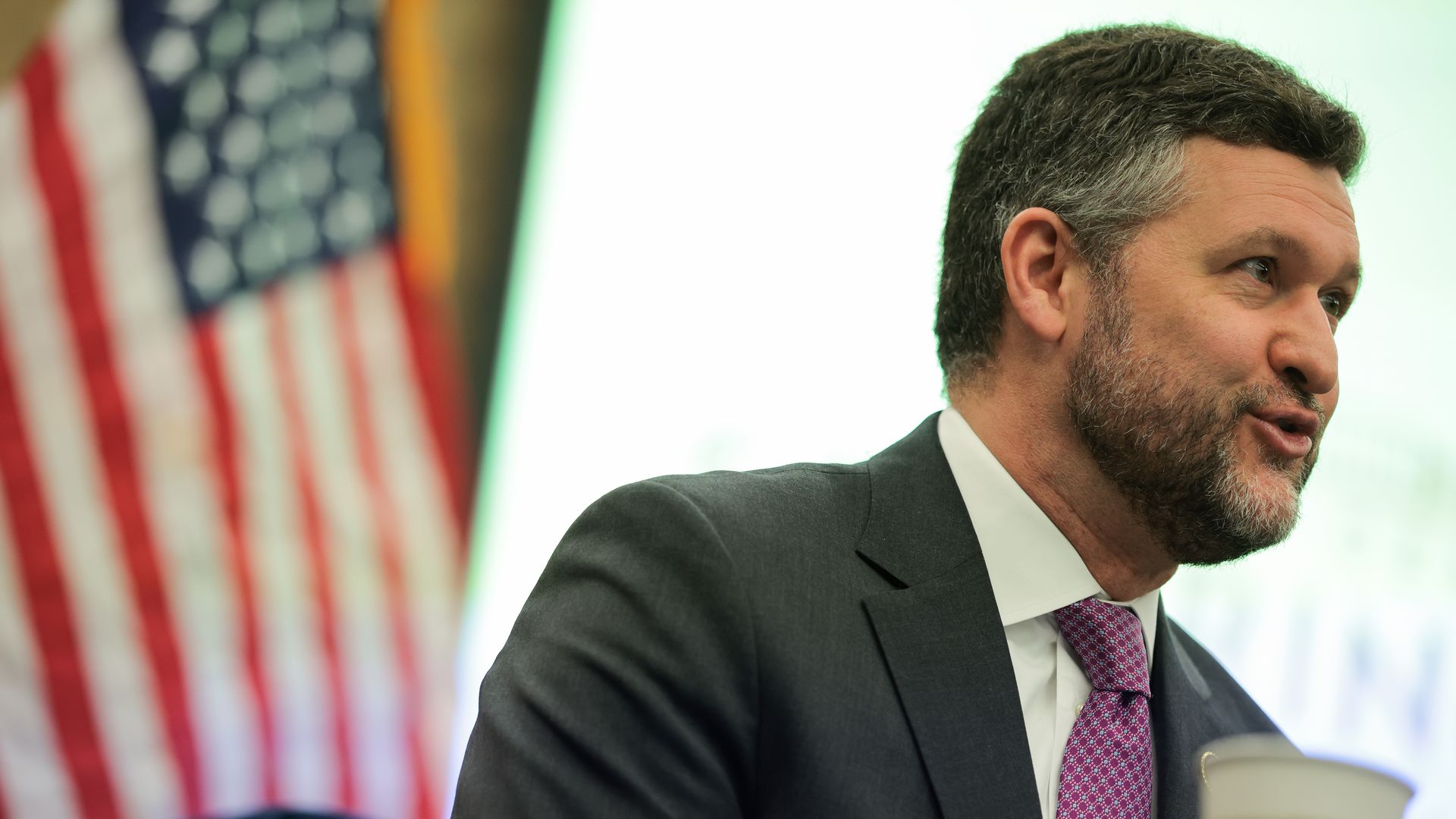 Man with dark hair and beard in a dark suit and purple tie speaking, with blurred American flag in background and a disposable coffee cup in foreground.