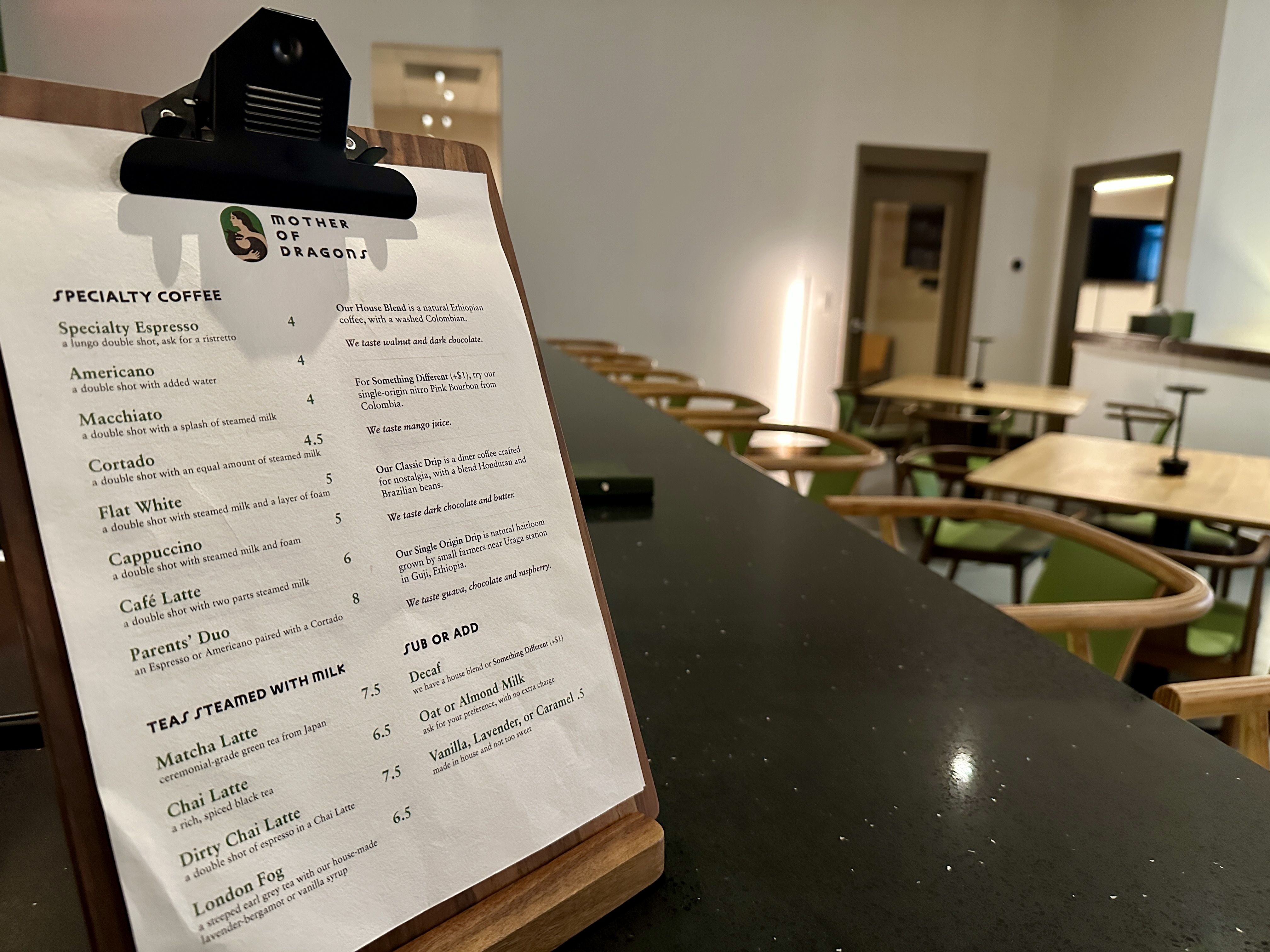 Cafe menu on clipboard at Mother of Dragons listing specialty coffees and teas, prices from 4 to 8.5, with modern seating and tables in a cozy cafe interior in background.