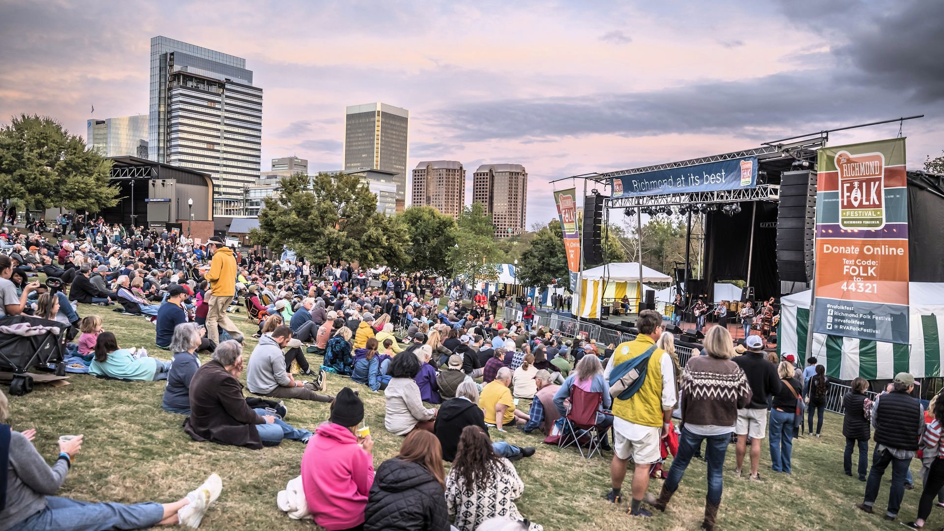 Large daytime crowd sits on a grassy park listening to a stage performance at Richmond Folk Festival; a city skyline with tall buildings looms in the background, banners and tents frame the stage.