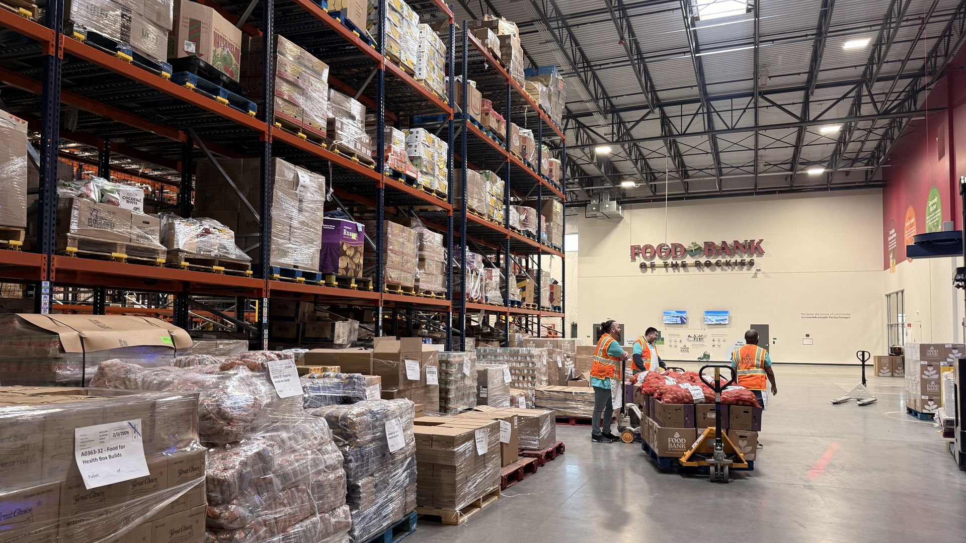 Interior of Food Bank of the Rockies warehouse with shelves stacked high with boxed goods and three workers in orange vests using pallet jacks to move boxes of food on pallets.