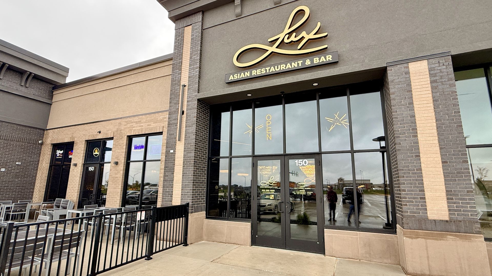 Exterior of a modern restaurant storefront with a gold "Lux" script sign and Asian Restaurant & Bar text on a gray wall. Brick pillars, large glass doors, outdoor seating to the left, and an overcast sky.