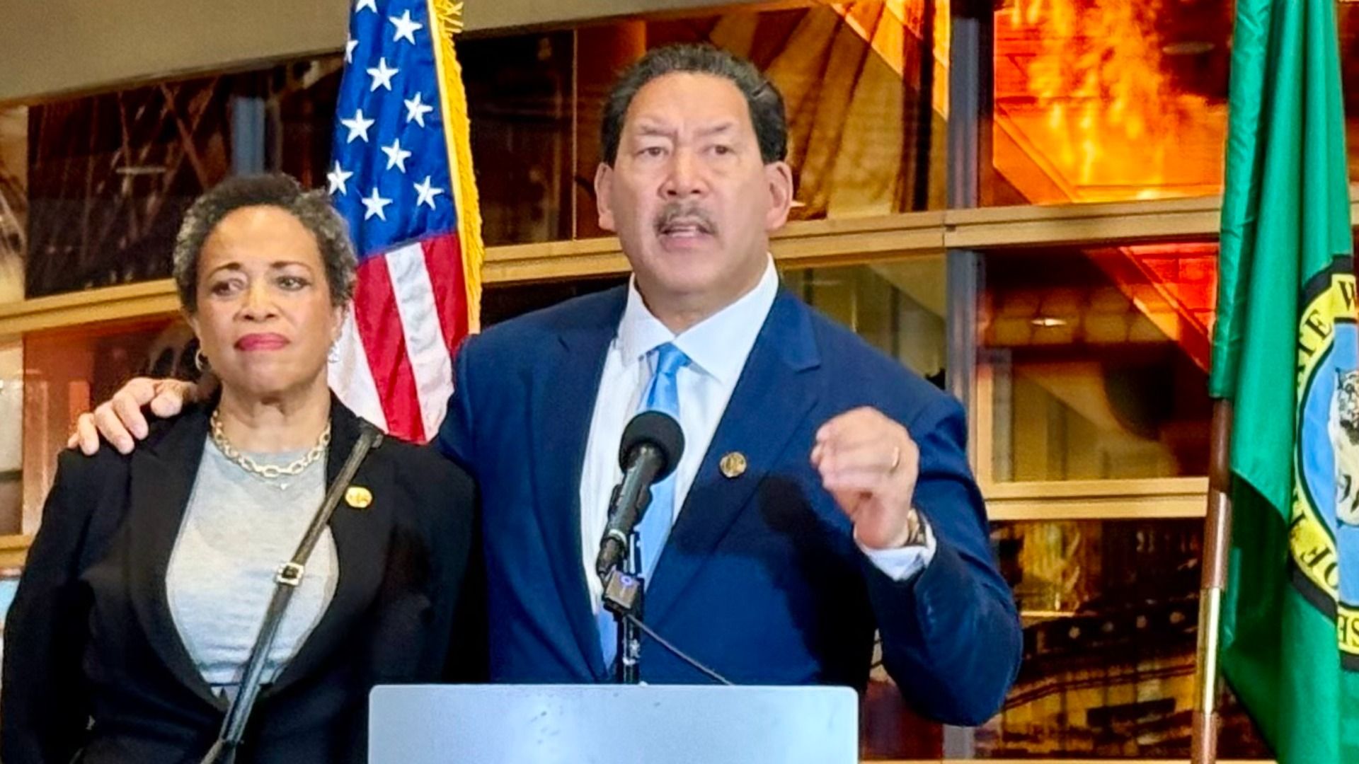 Two people, Bruce Harrell and Joanne Harrell, stand at a podium with microphones; Harrell is in a blue suit and tie gestures while Joanne, in a black blazer, looks on. U.S. and Washington state flags are behind them.