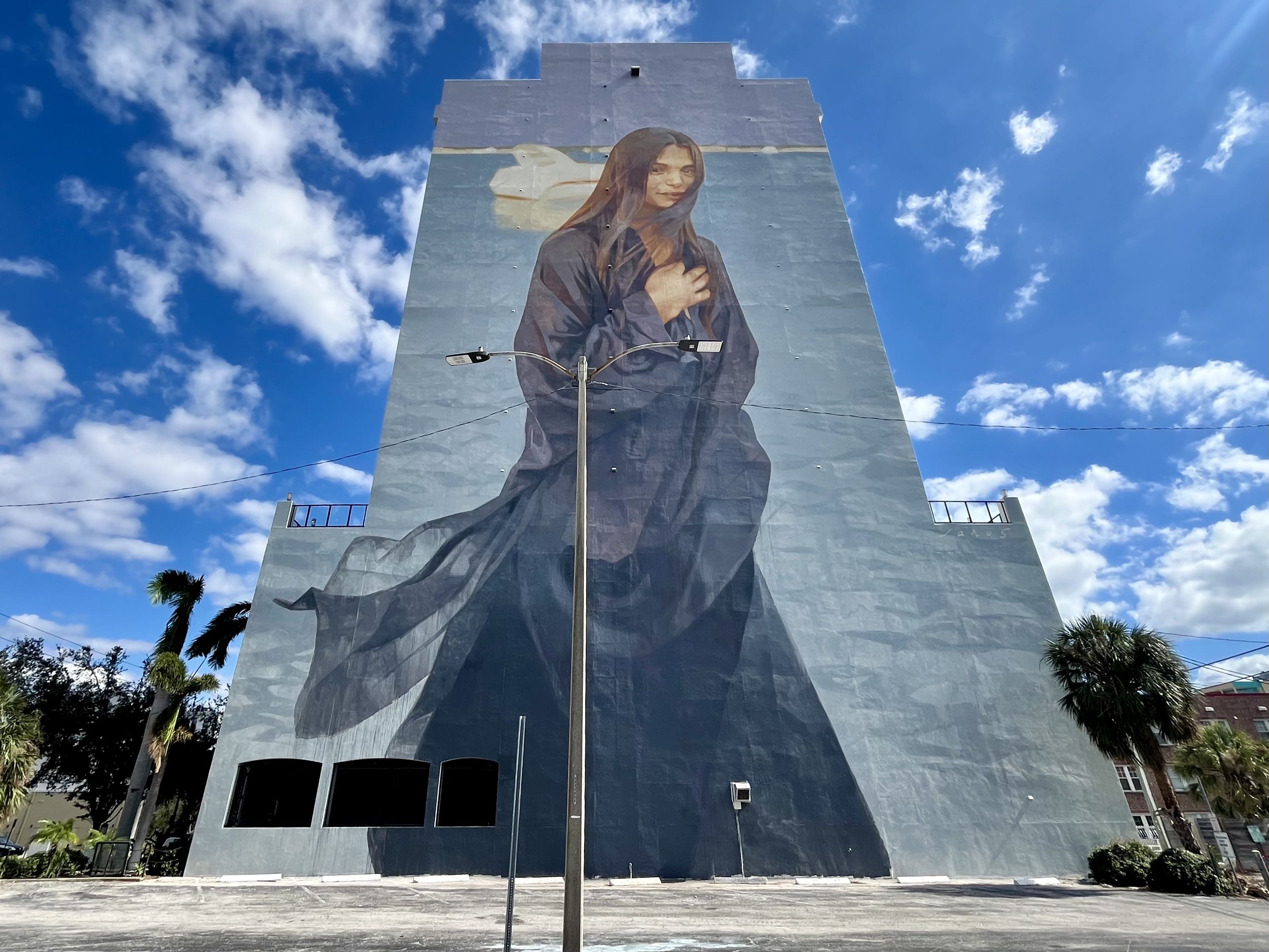 A mural painted on super tall light blue wall showing a woman in a black cloak smiling coyly at the viewer.