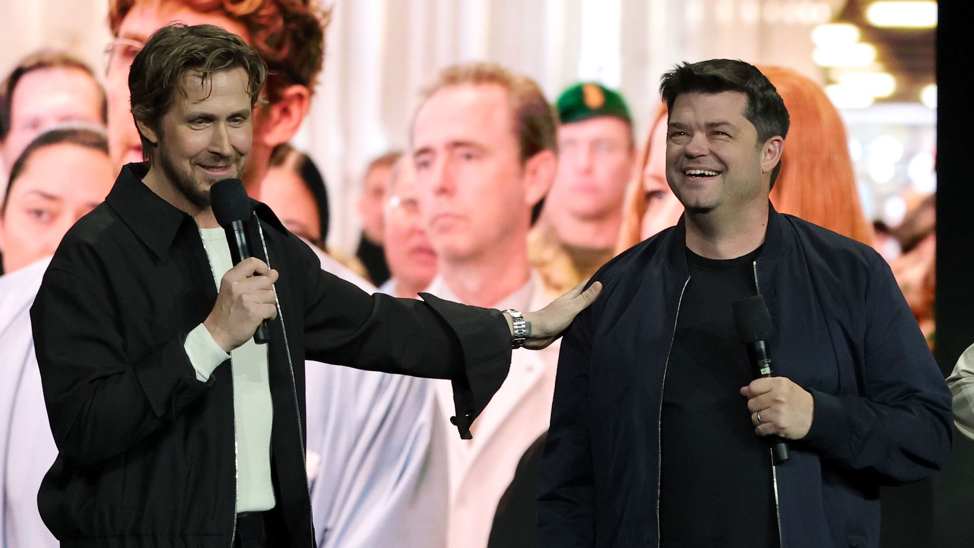Ryan Gosling speaks into a mike while putting his hand on the shoulder of Christopher Miller. They wear black jackets and stand in front of a screen with a scene from a film.
