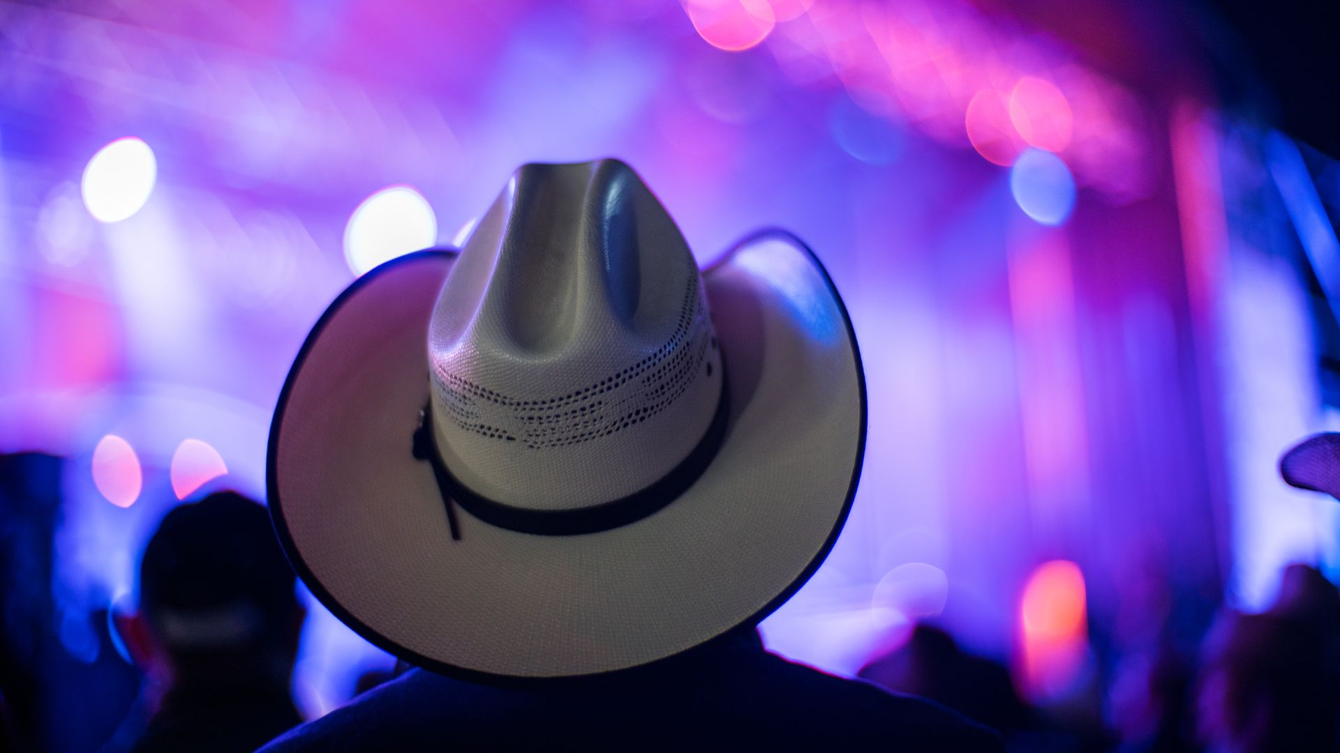 A photo taken from behind a person wearing a cowboy hat in front of a stage with blurred out blue lights