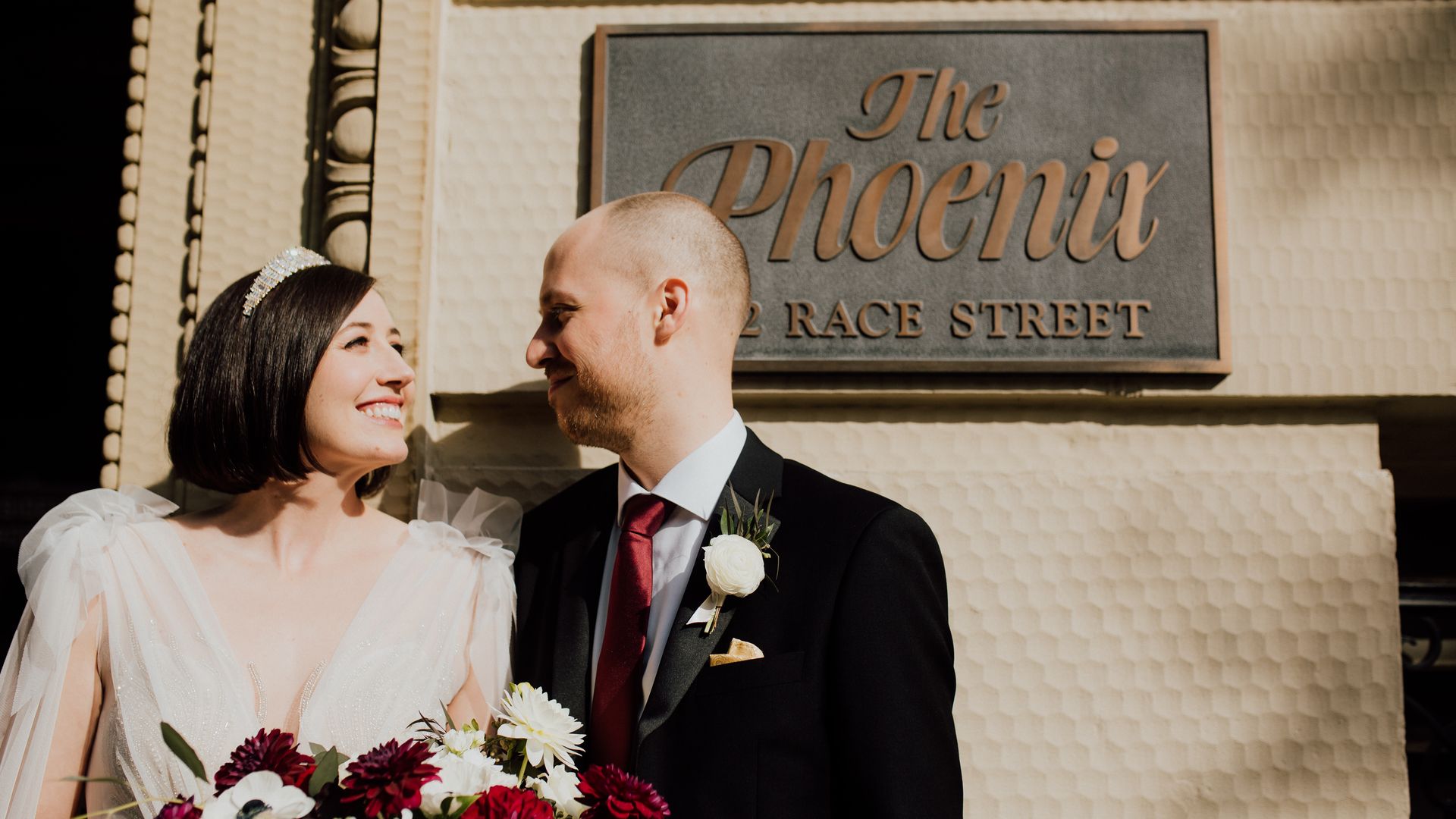 Joyful bride and groom in wedding attire smile at each other in front of a beige tiled wall beside a sign reading "The Phoenix" in elegant script; the bride holds a red-and-white bouquet.