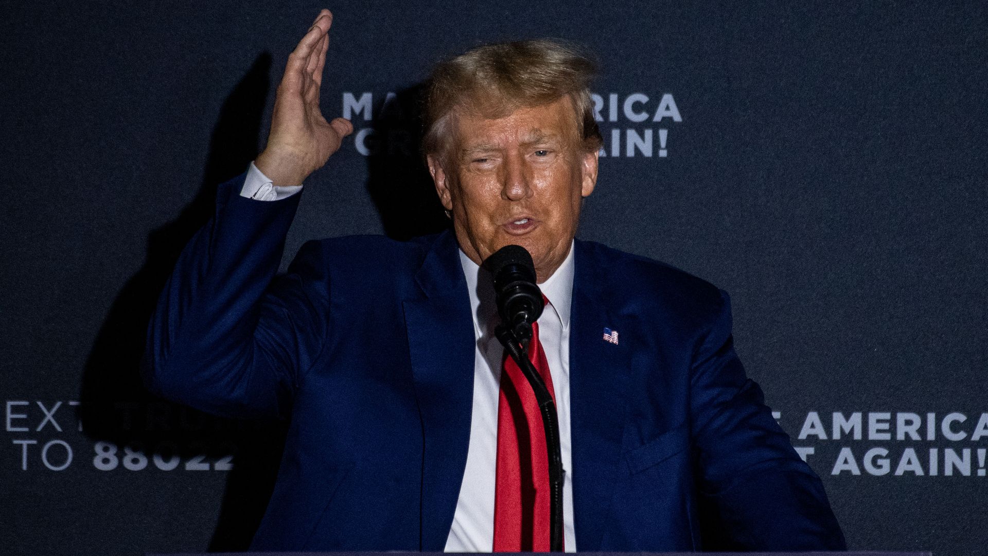 Trump speaking at a rally in front of a backdrop that says "Make America Great Again!" He wears a blue suit and red tie. One of his arms is in the air as he speaks.