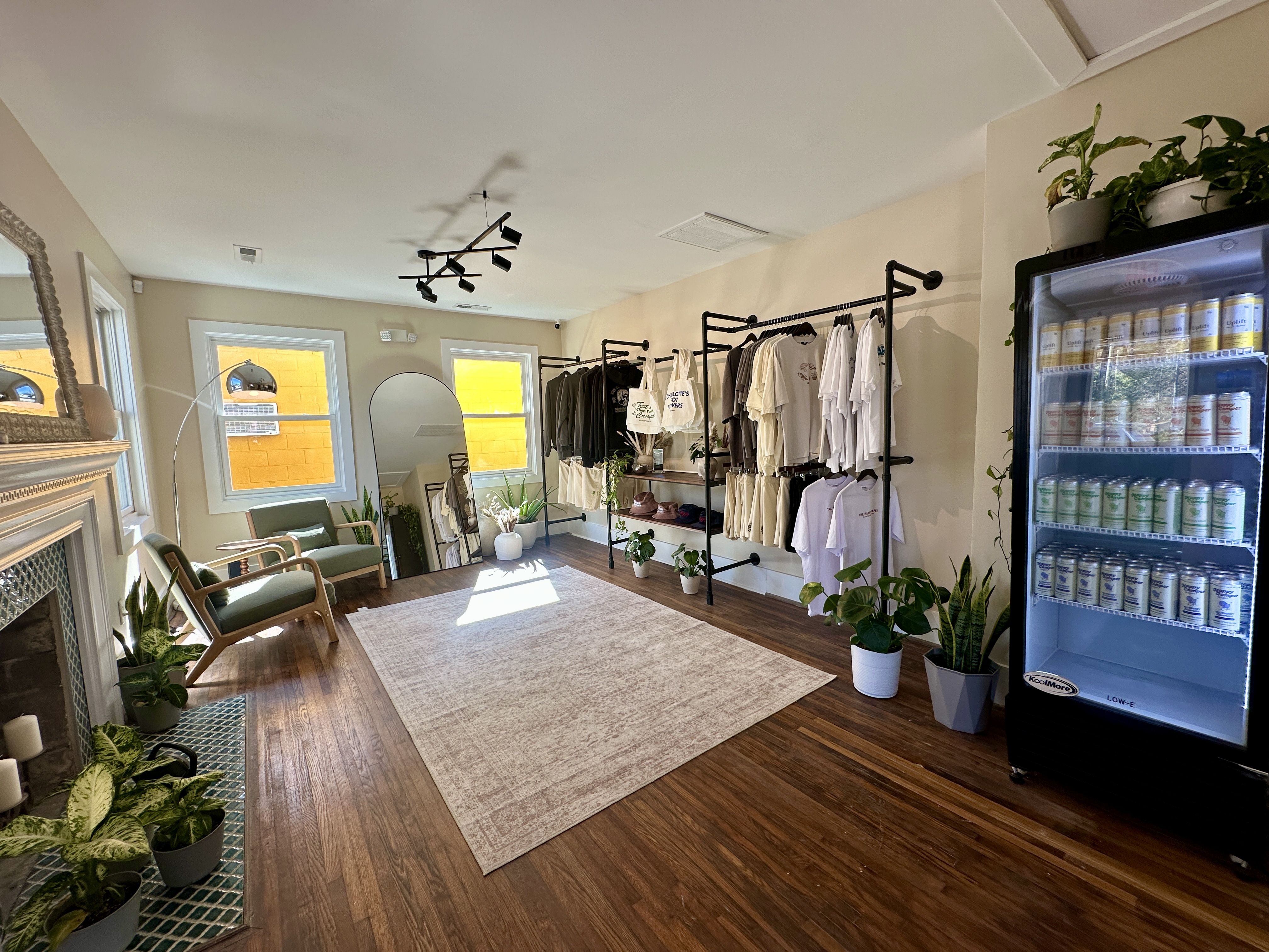 Sunlit boutique with warm wooden floors, a large rug, and clothing racks along the right wall. Green plants near a circular mirror and two chairs by the left window; a drinks fridge on the right.