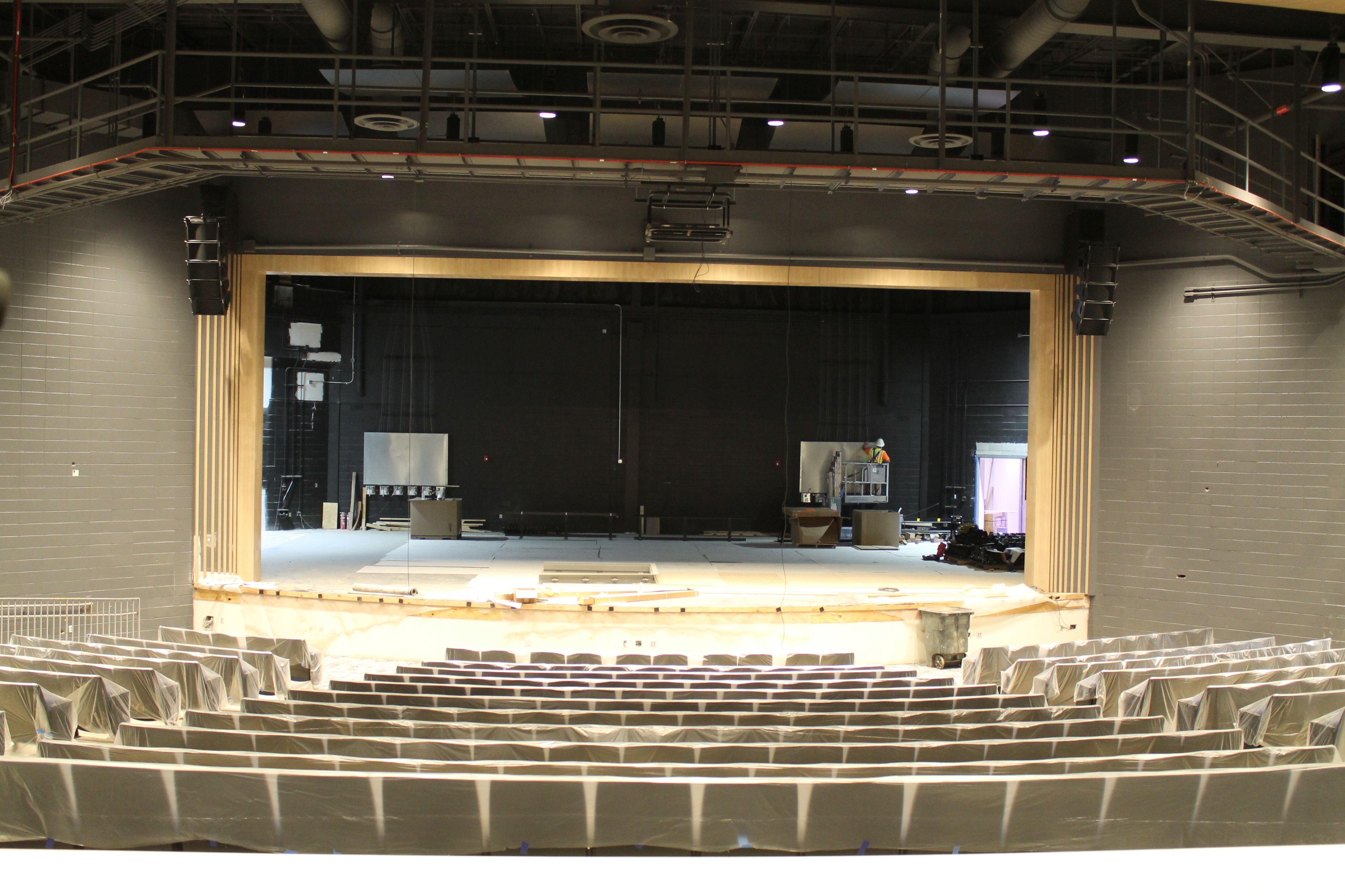 Indoor theater under construction: rows of plastic-covered seats face a stage with a wooden proscenium, scaffolding and hanging cables overhead; workers in hard hats near back of stage.