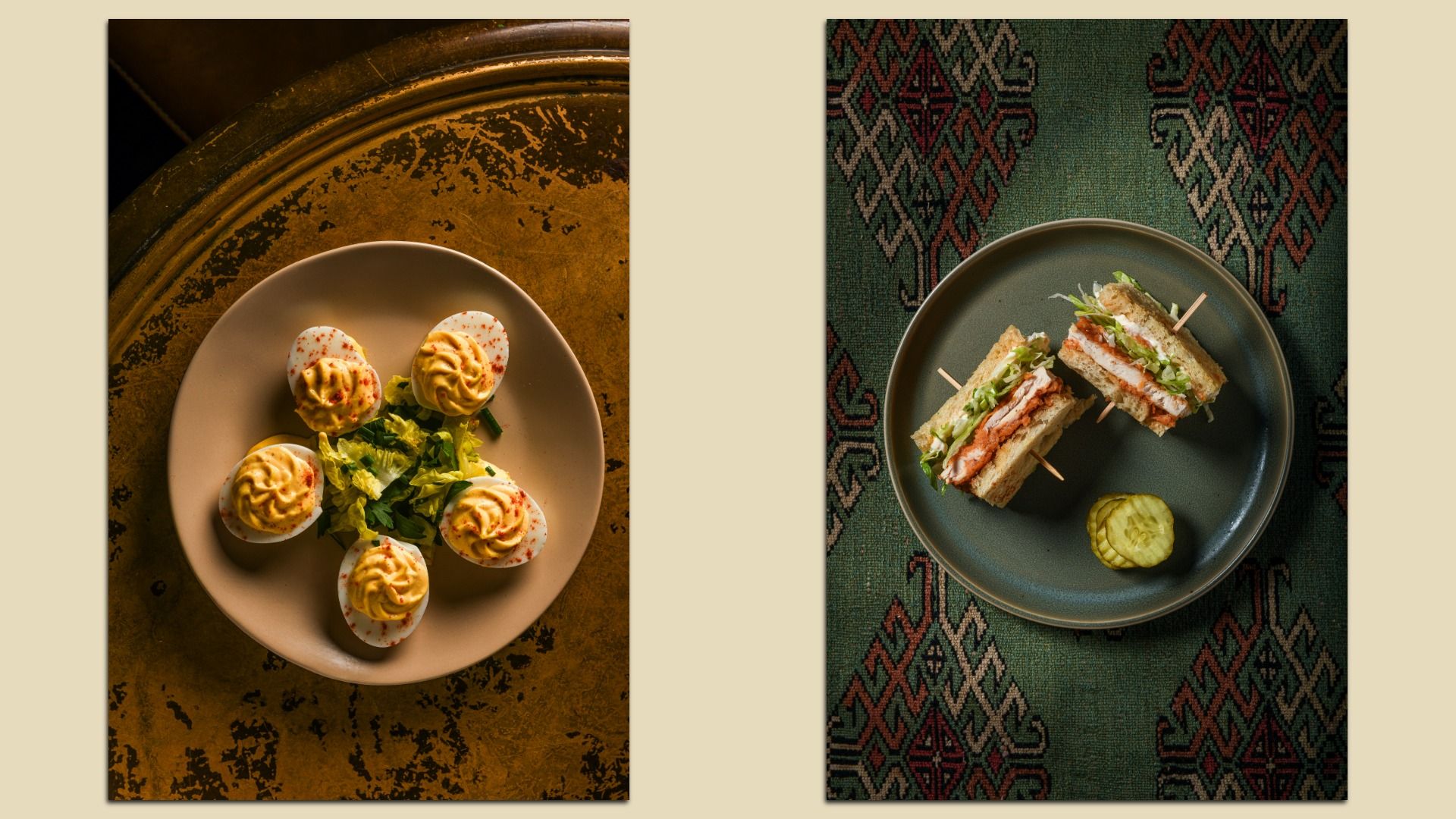 Two plates of food: one with five deviled eggs garnished with paprika and greens on a bronze surface, the other with two sandwich halves and pickle slices on a dark plate over a patterned green cloth.