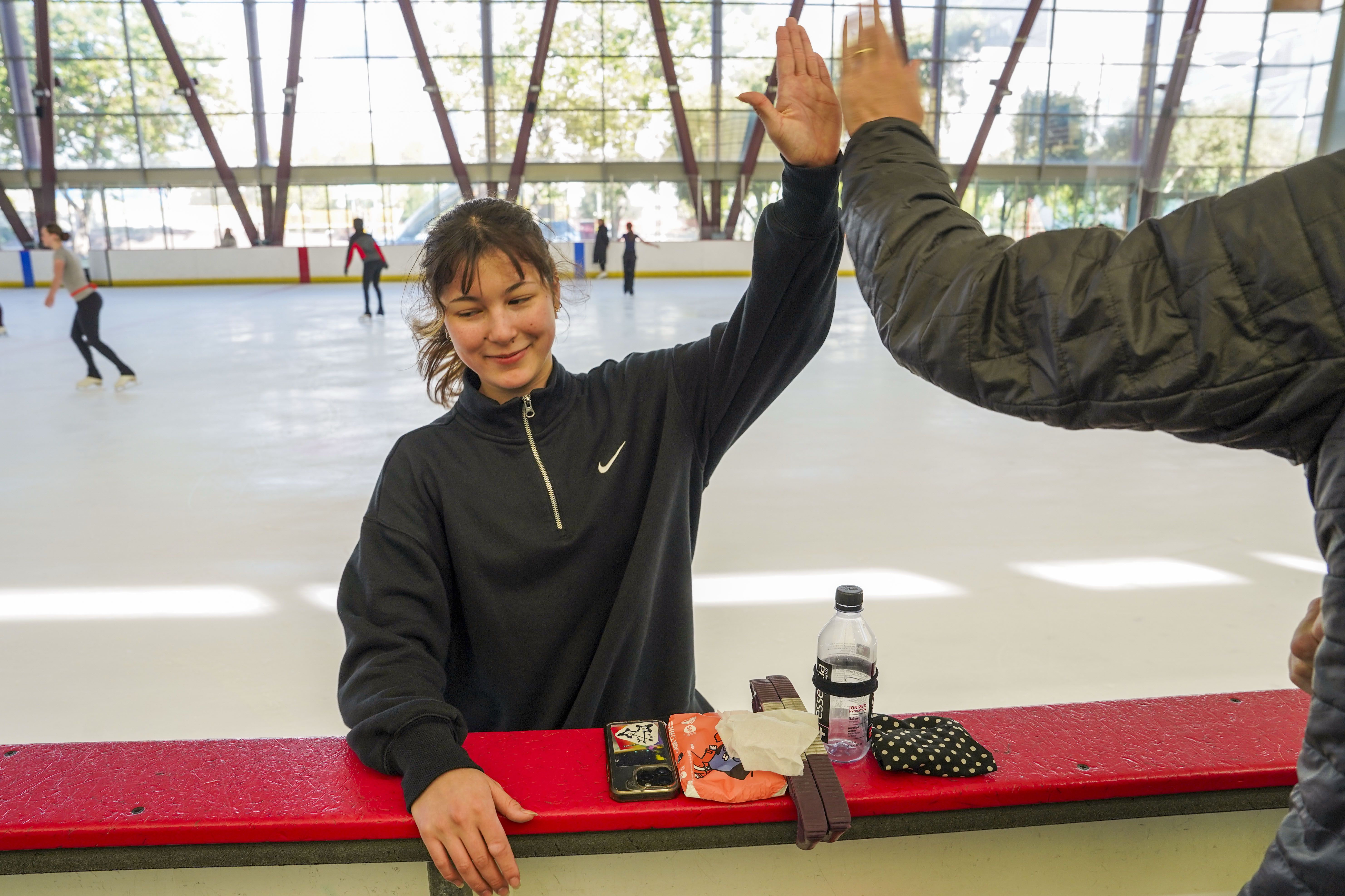 Smiling young woman in black Nike sweatshirt giving a high five at an indoor ice skating rink, with ice skaters in the background and personal items on the red barrier.
