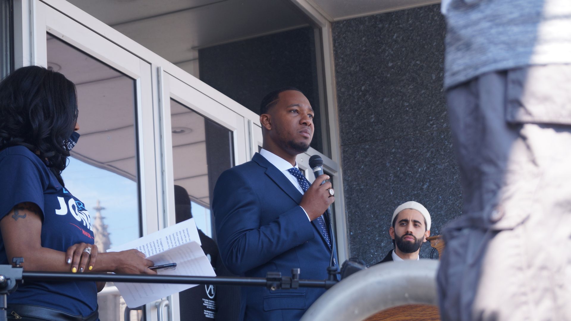 Black man in suit holding microphone outside doorway