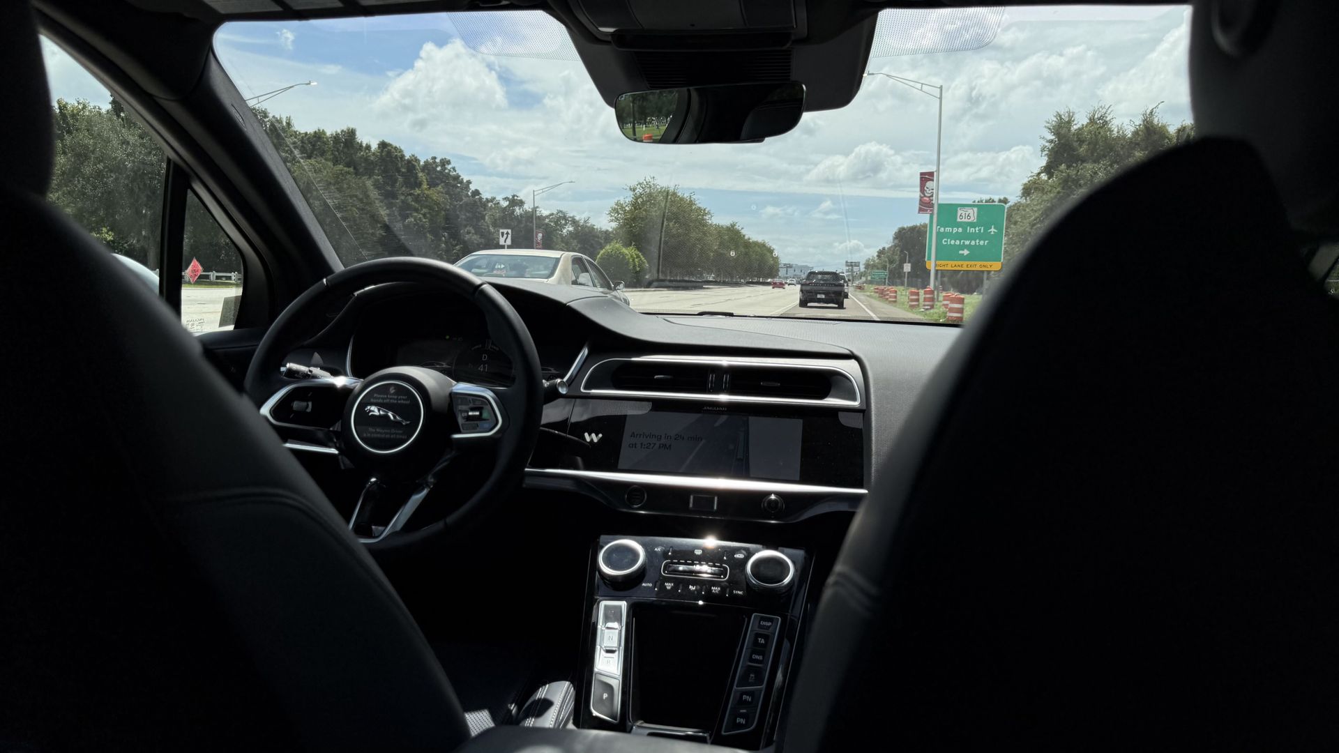 View from the back seat of a car: black steering wheel, glossy center console, and dashboard. Through the windshield, a sunny highway with trees and a green exit sign for Tampa Intl and Clearwater.