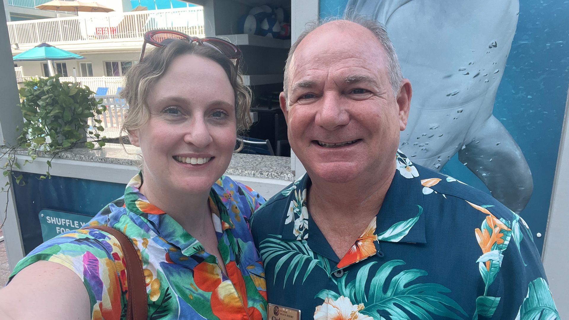 Smiling man and woman wearing colorful floral shirts, standing outdoors near a pool area with umbrellas and a large shark image in the background.