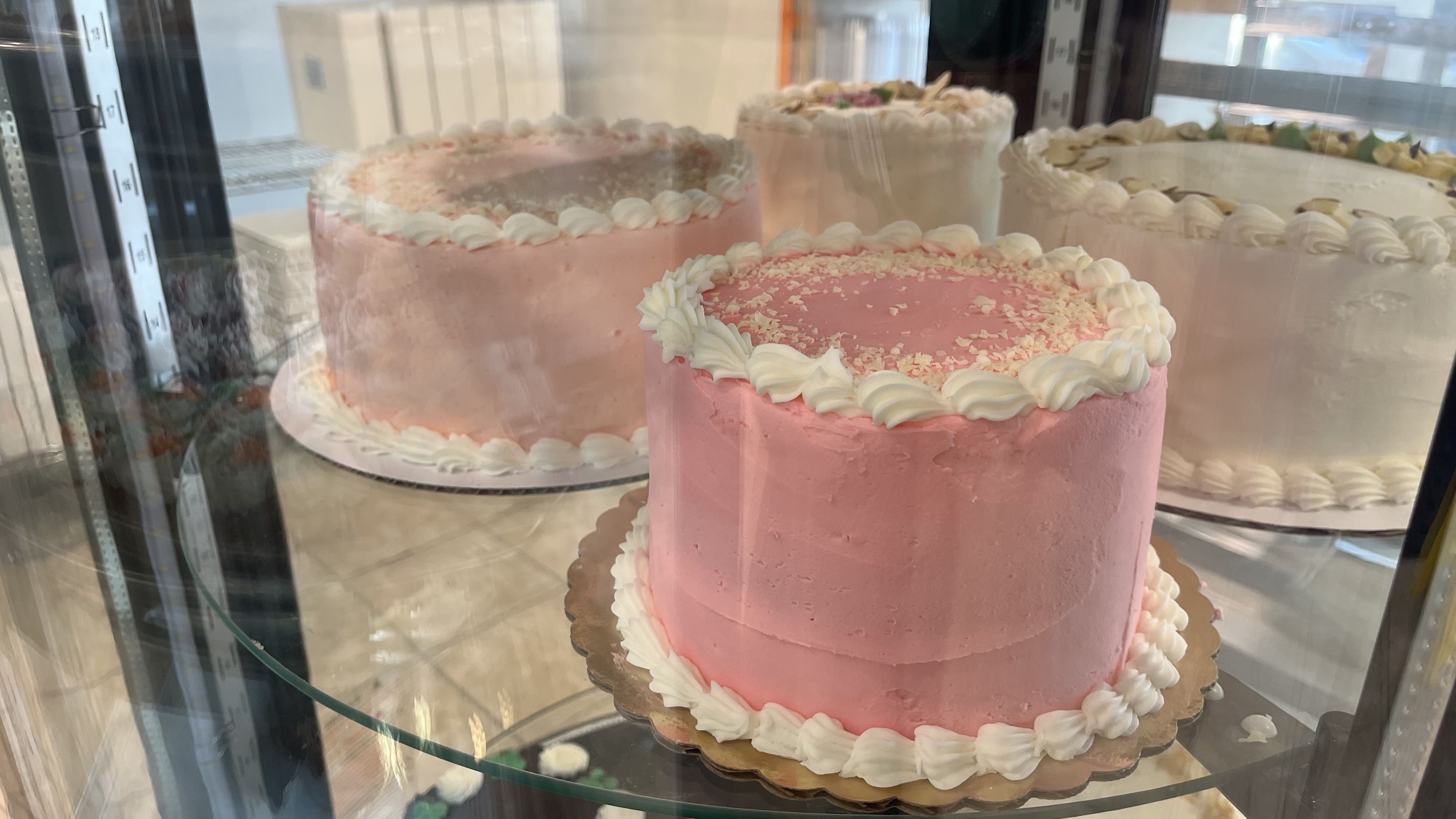 Pink frosted cake with white piped border on a gold stand inside a bakery display case; other cakes with white frosting visible in the background.