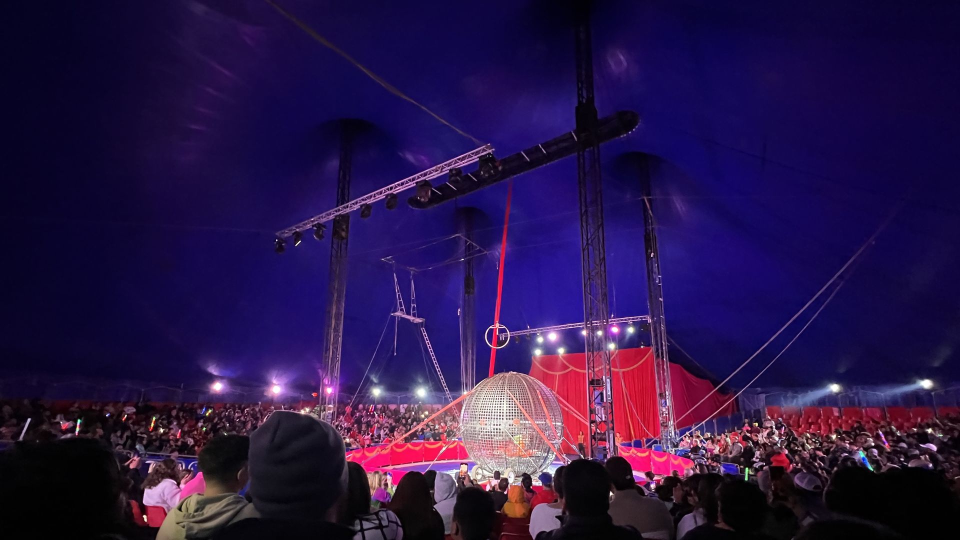 A picture inside a round tent with at least a hundred people facing a stage where there's a metallic round cage and a motorcyclist inside of it.