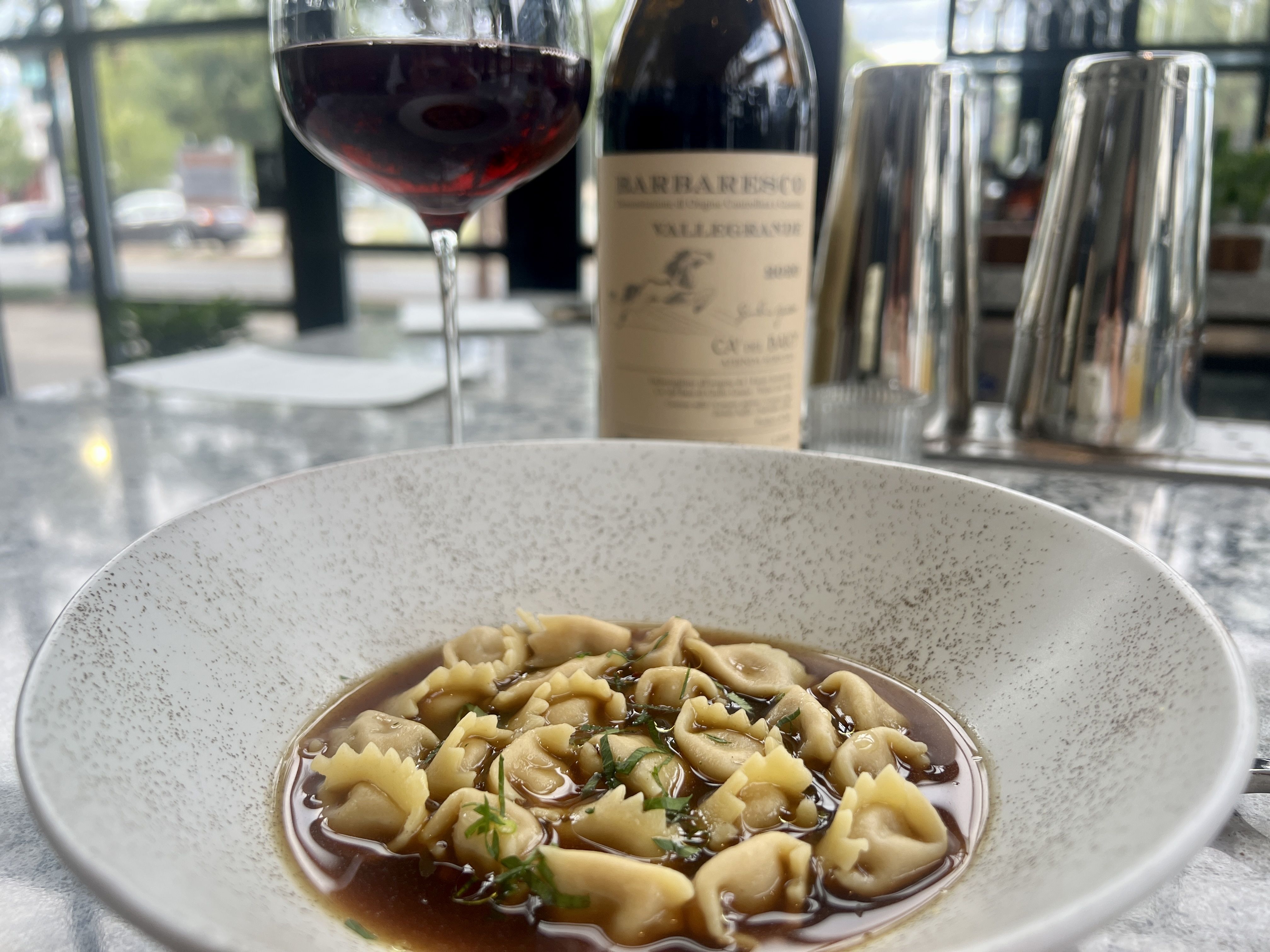 Close-up of a speckled bowl of pasta in brown sauce with herbs. In the blurred background, a glass of red wine and a Barbaresco bottle on a restaurant table by a window.