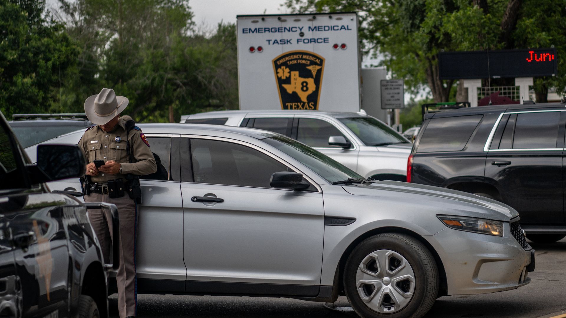 A law enforcement officer stands in front of a car looking down at his phone