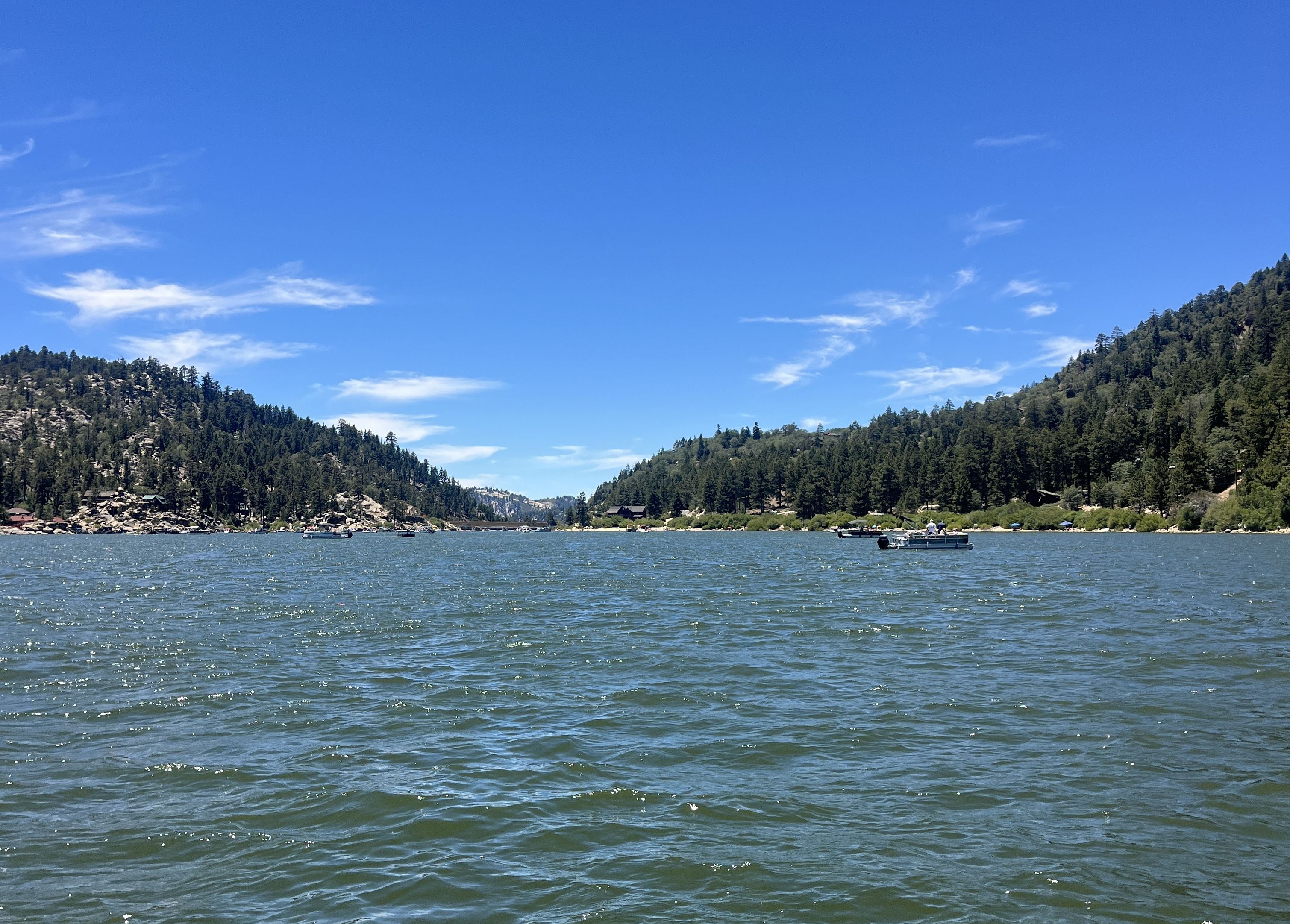 Wide blue sky over a calm lake, with forested hills on both shores and small boats near the rocky shoreline.