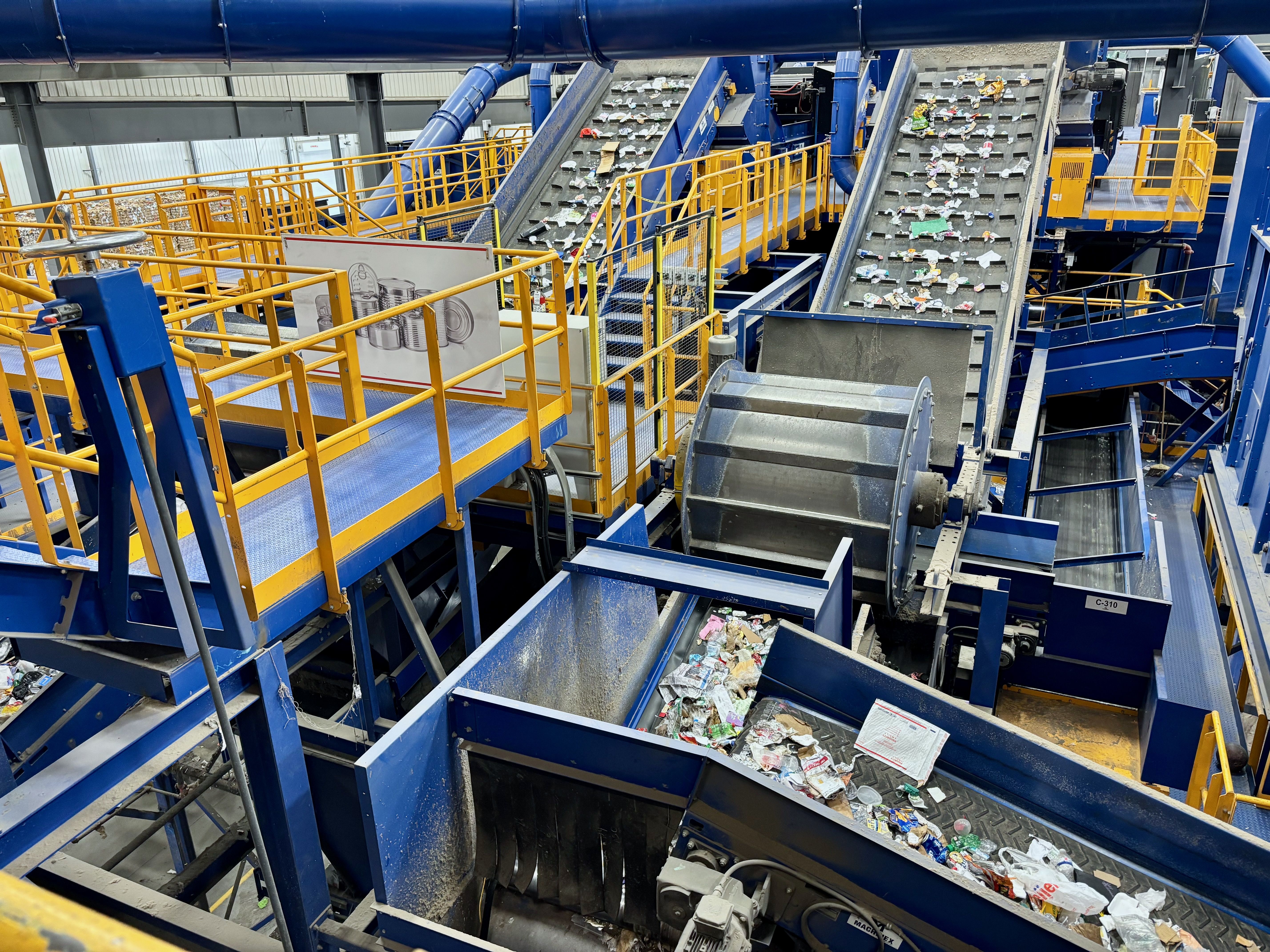 Industrial recycling facility with blue and yellow machinery. Conveyor belts move scraps toward a metal chute, flanked by yellow railings and blue structures.