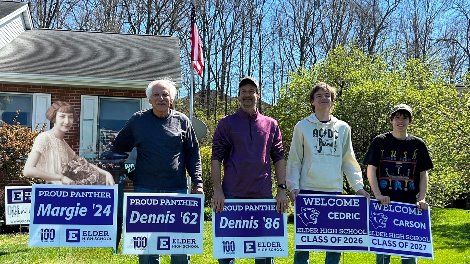 Five people stand on a sunny lawn in front of a brick house with an American flag, holding blue Elder High School signs; a life-size cutout of a girl sits at the left.