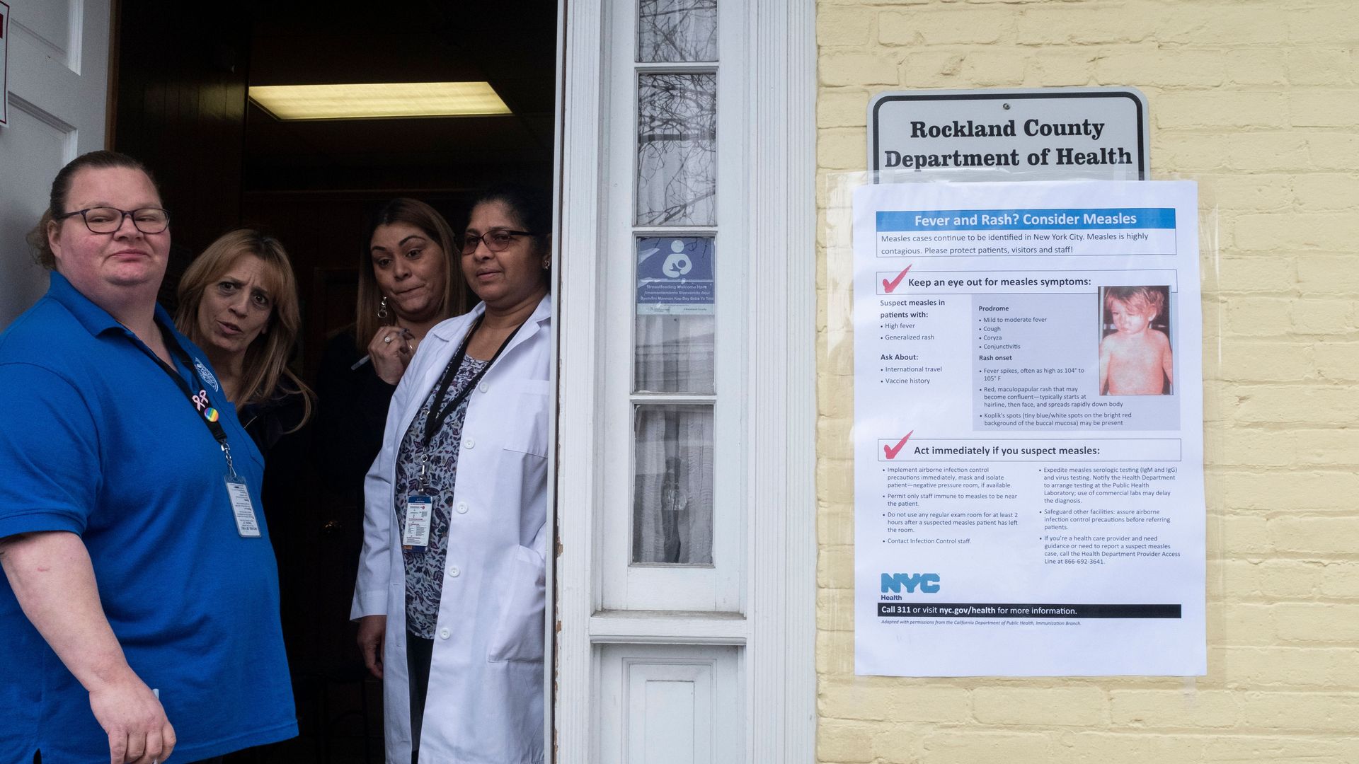 Photo of nurses at a clinic in Rockland County, NY, peering outside the door next to a measles information sign