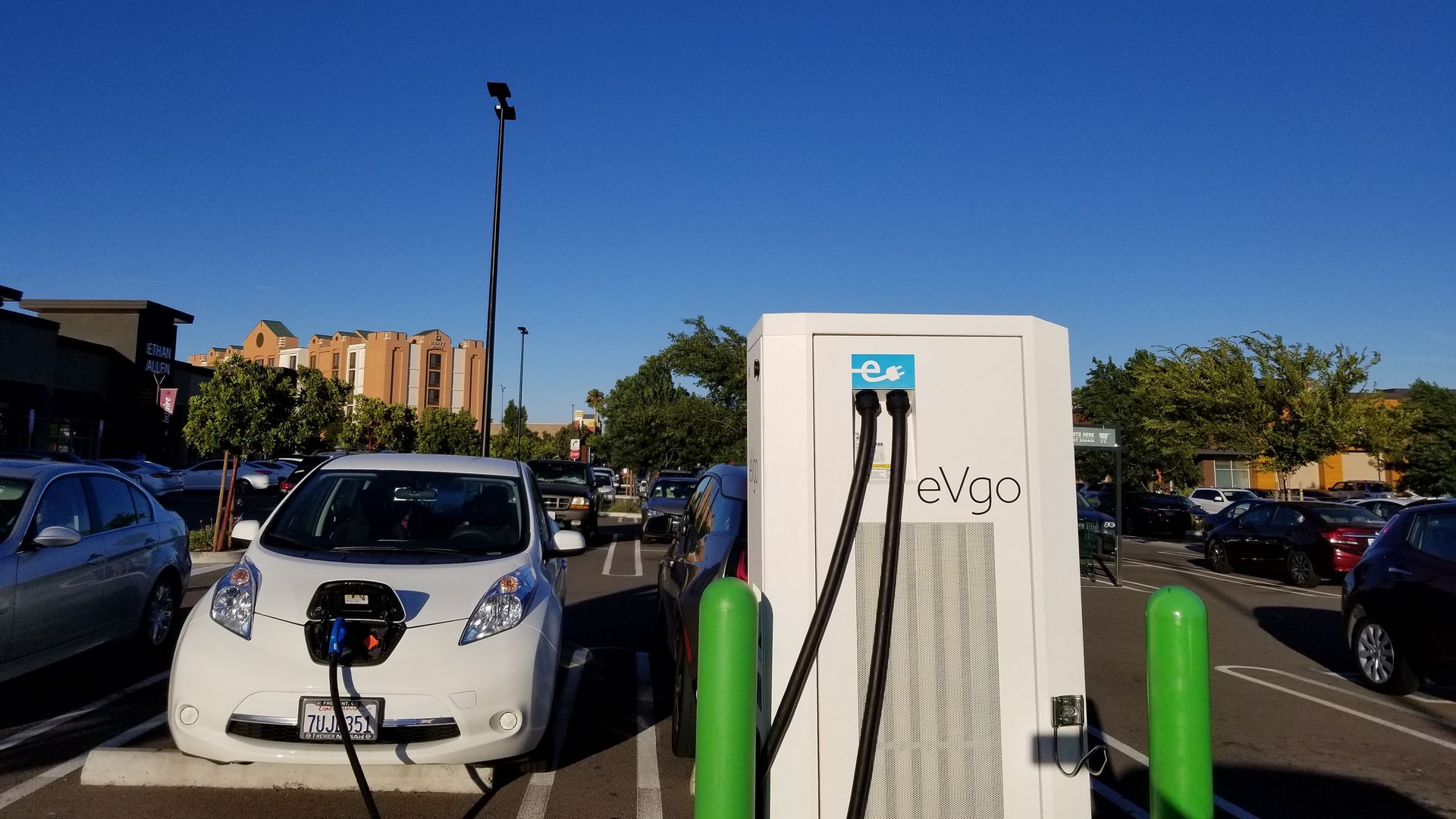 In this image, an electric car is plugged into a charger in a sunny parking lot