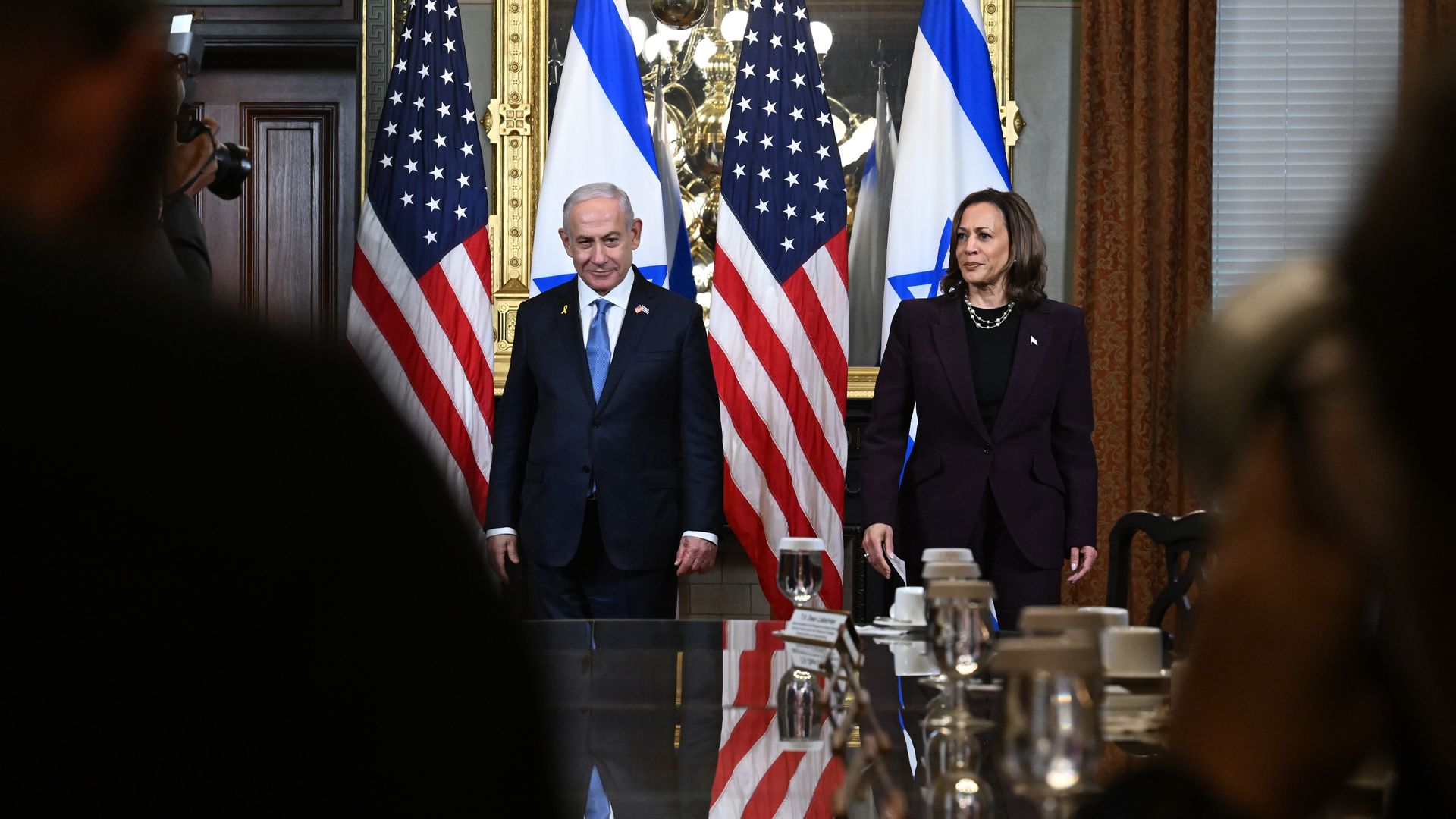 Benjamin Netanyahu wearing a blue suit and Kamala Harris wearing a purple suit, standing in front of American and Israeli flags.