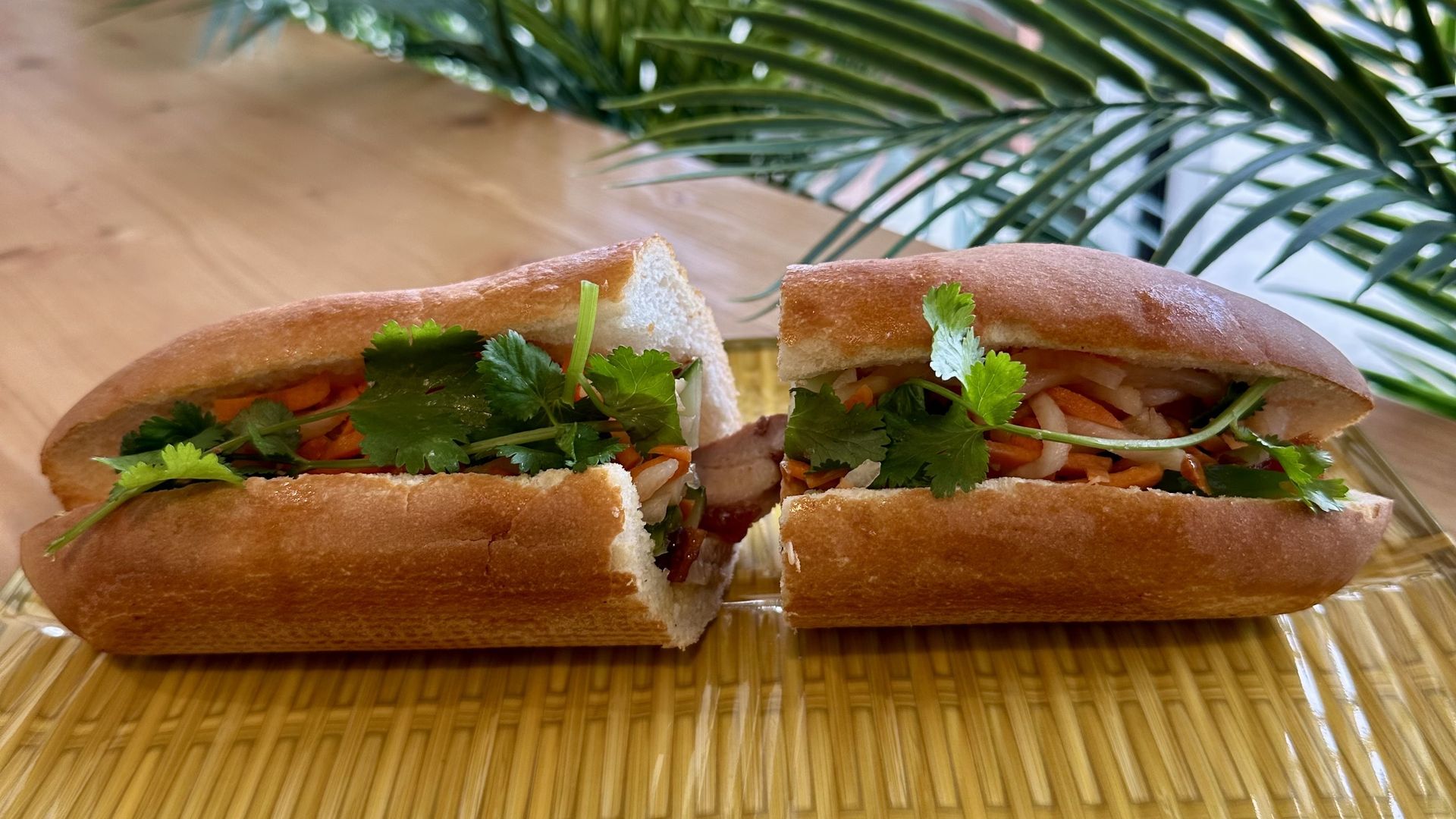 Two halves of a baguette sandwich filled with cilantro and vegetables sit on a woven placemat, with blurred green plants and a windowed, bright indoor background.
