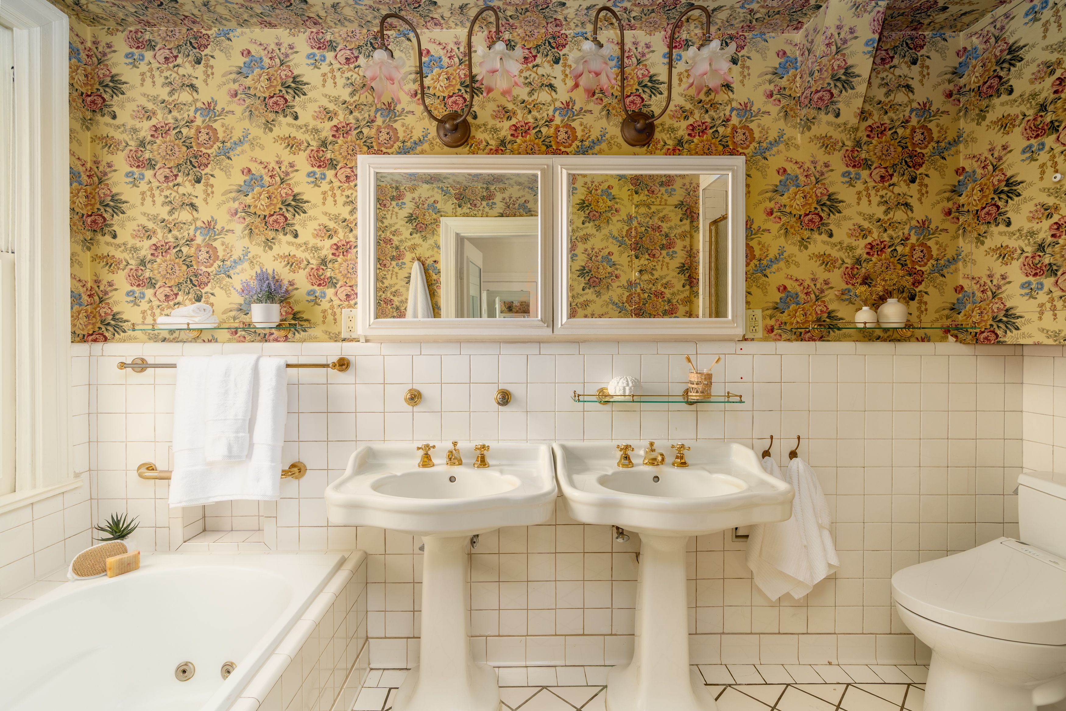 Vintage bathroom with yellow floral wallpaper, twin white pedestal sinks, gold faucets, large framed mirrors, frosted pink flower lights, white tiled walls, towels on gold bars, and a bathtub on the left.