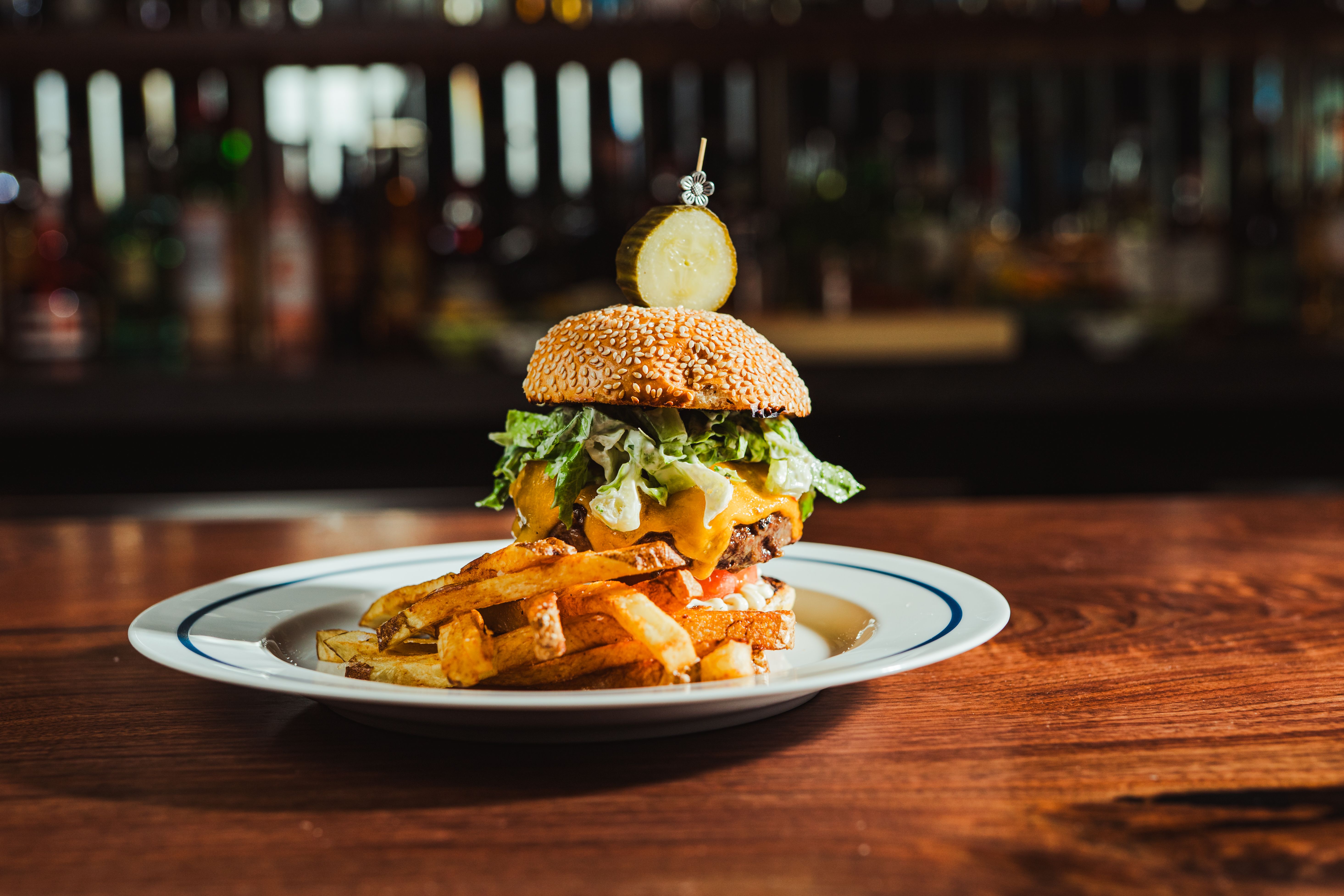 Stacked cheeseburger with sesame bun, lettuce, cheese, and sauce on fries; topped with a pickle slice on a decorative skewer, against a blurred bar backdrop.